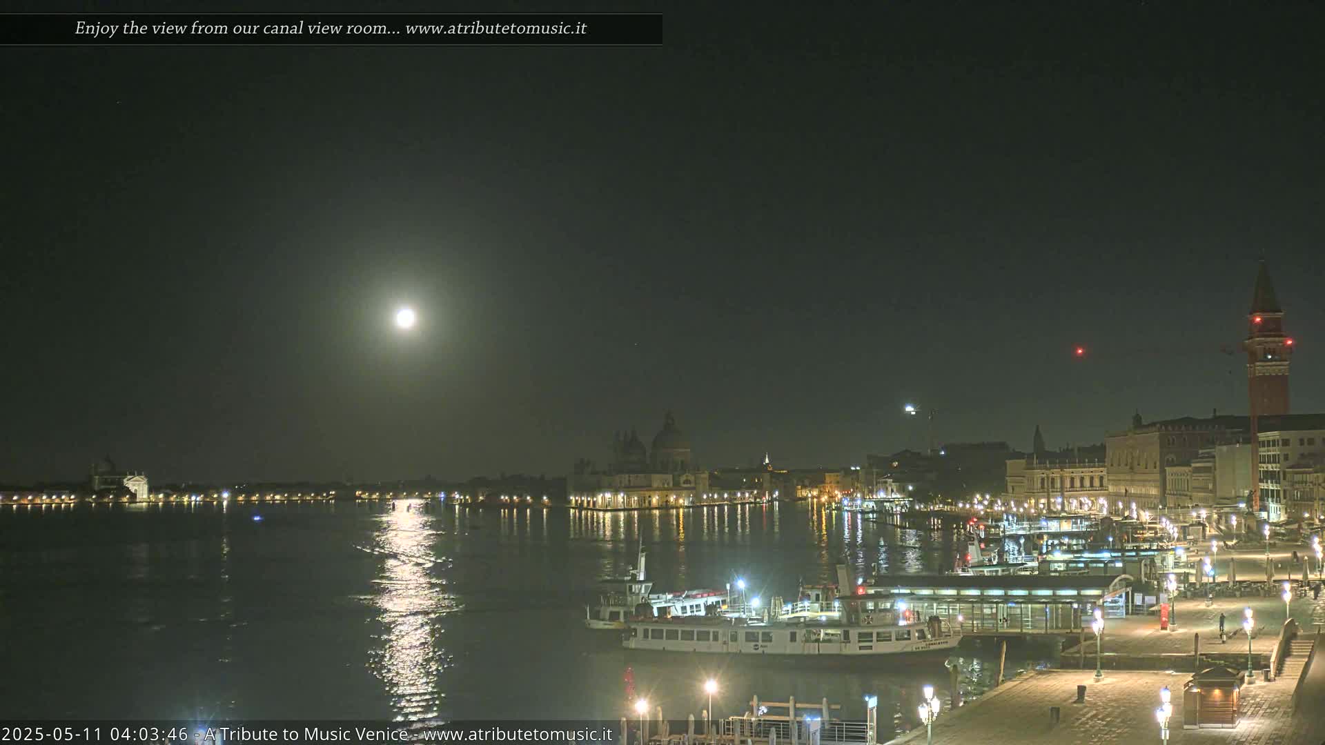 A brightly lit cityscape at night, under a full moon, is reflected on calm water with boats moored along a waterfront.