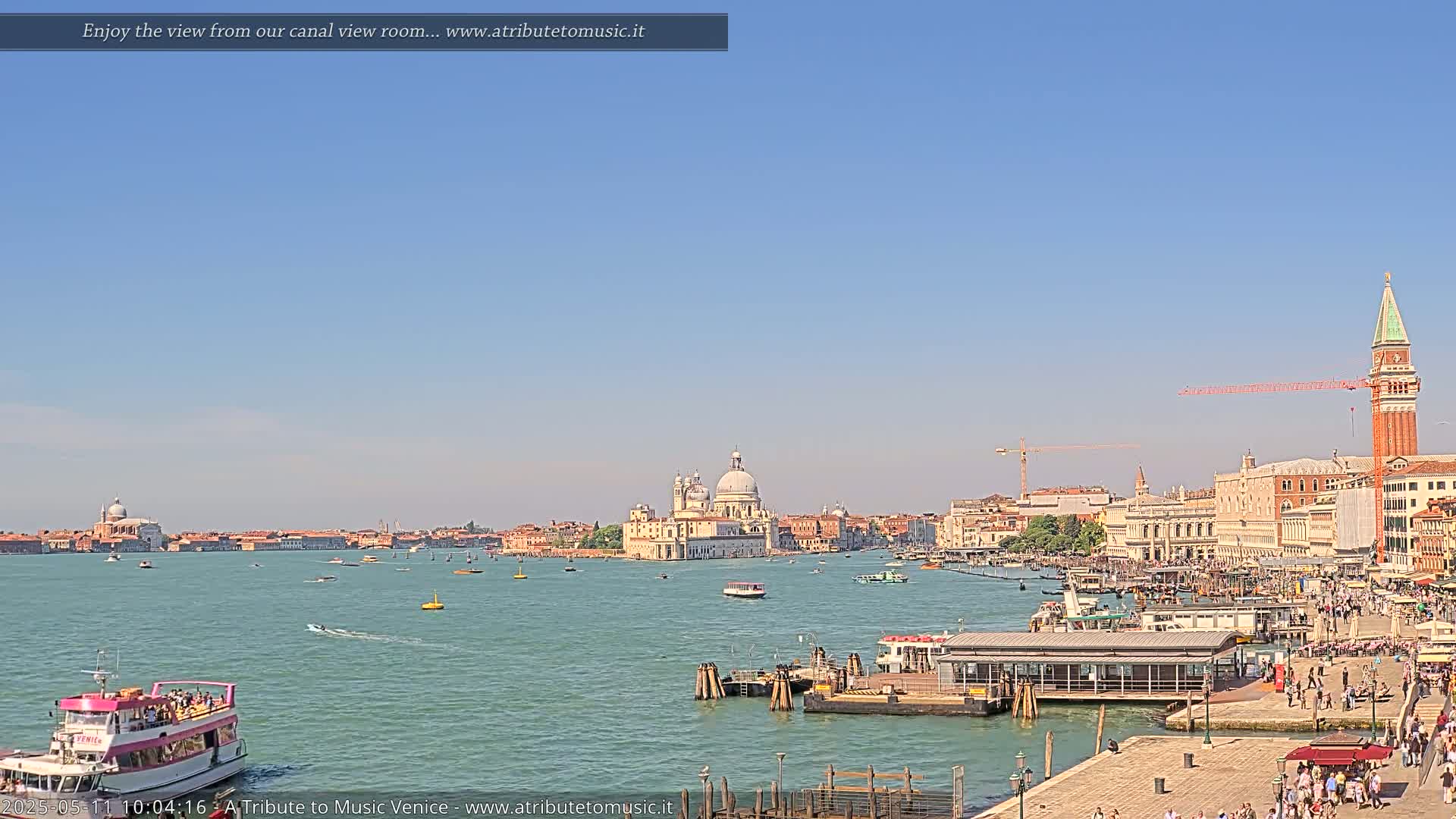 A sunny day in Venice shows a canal with boats, a waterfront with people, and a distant view of buildings including a large domed church and a tall bell tower.