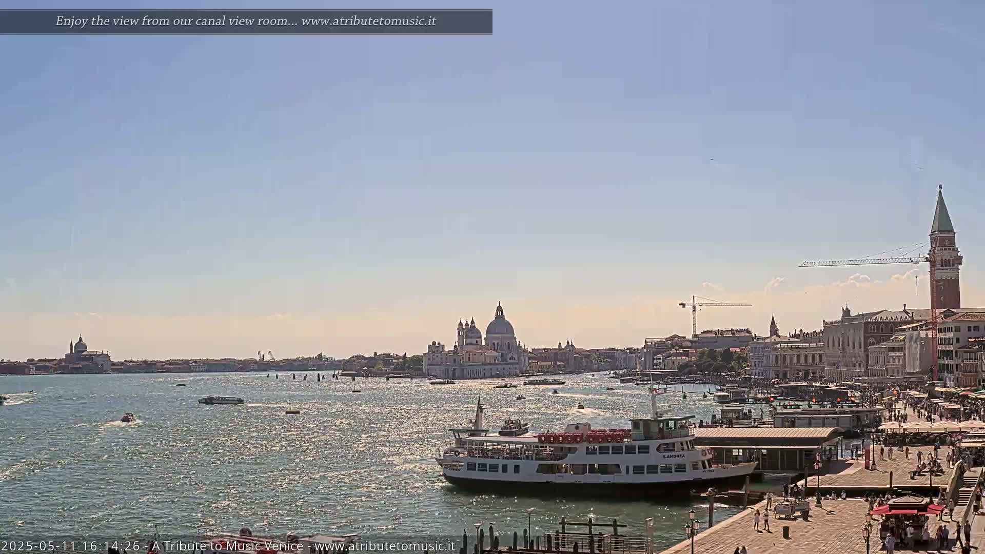 A sunny day in Venice shows a wide canal with a passenger ferry and several smaller boats, and the city skyline including the Basilica di Santa Maria della Salute in the distance.