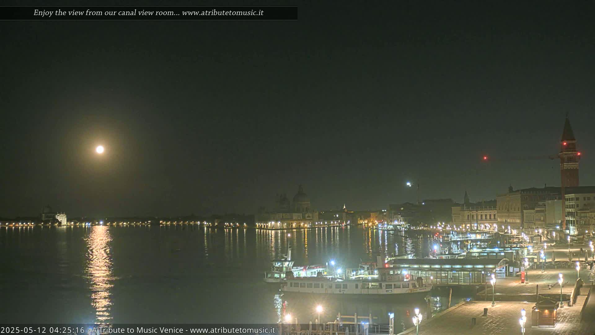 A bright moon illuminates a nighttime view of a canal, boats, and buildings along a waterfront, under a clear, dark sky.