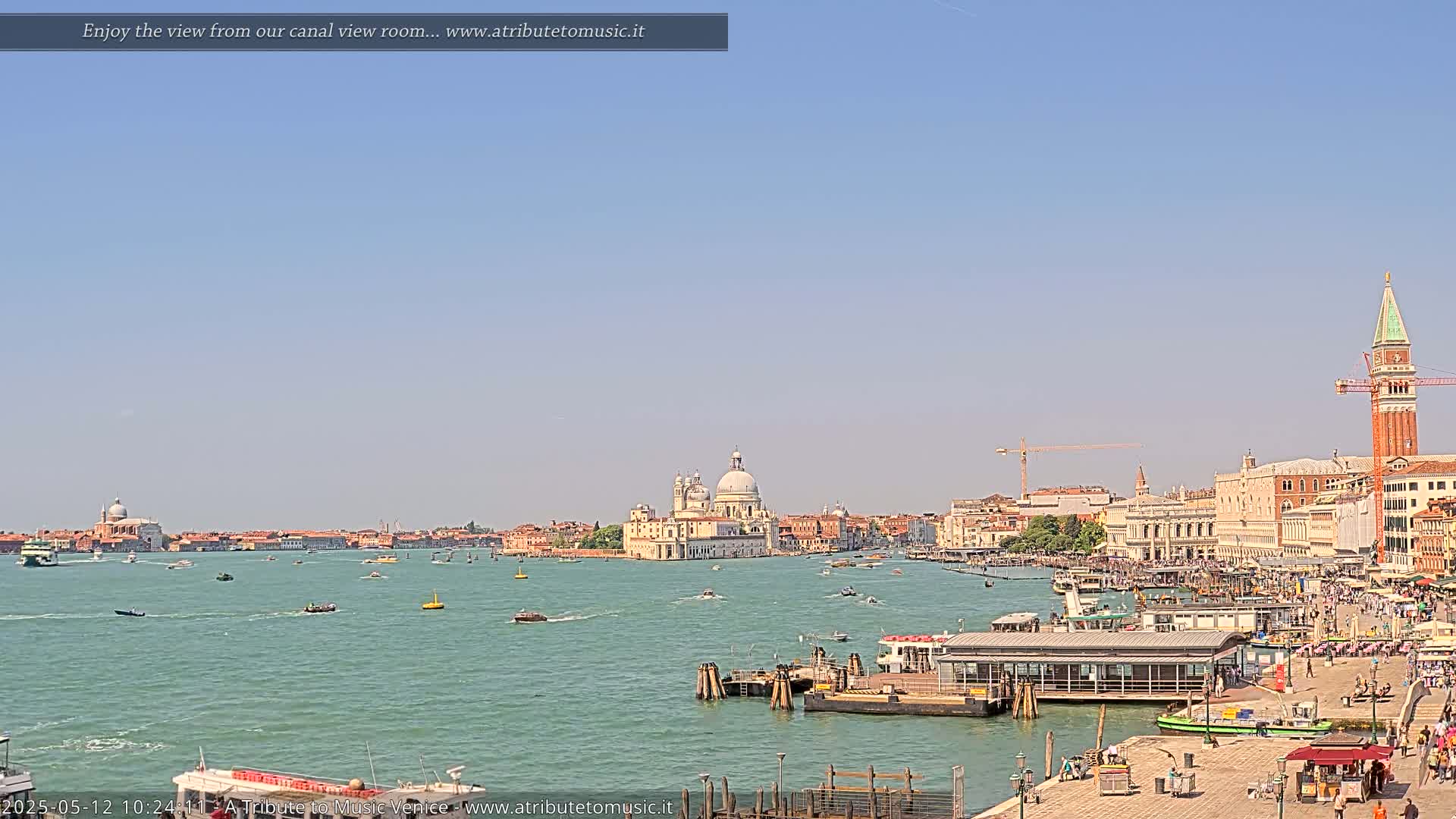 A sunny day in Venice shows a wide canal with boats, buildings lining the waterfront, and a large church in the distance.