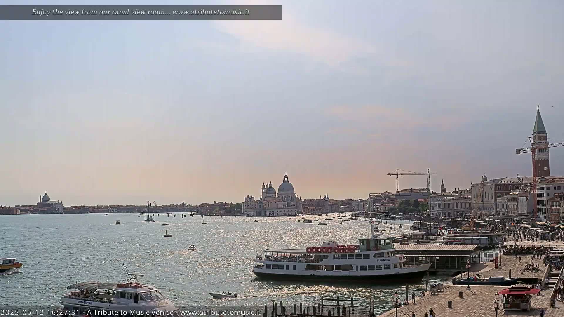 A sunny Venetian canal scene shows several boats on the water, with a large passenger ferry docked near a waterfront building, and the Basilica di Santa Maria della Salute visible in the distance.