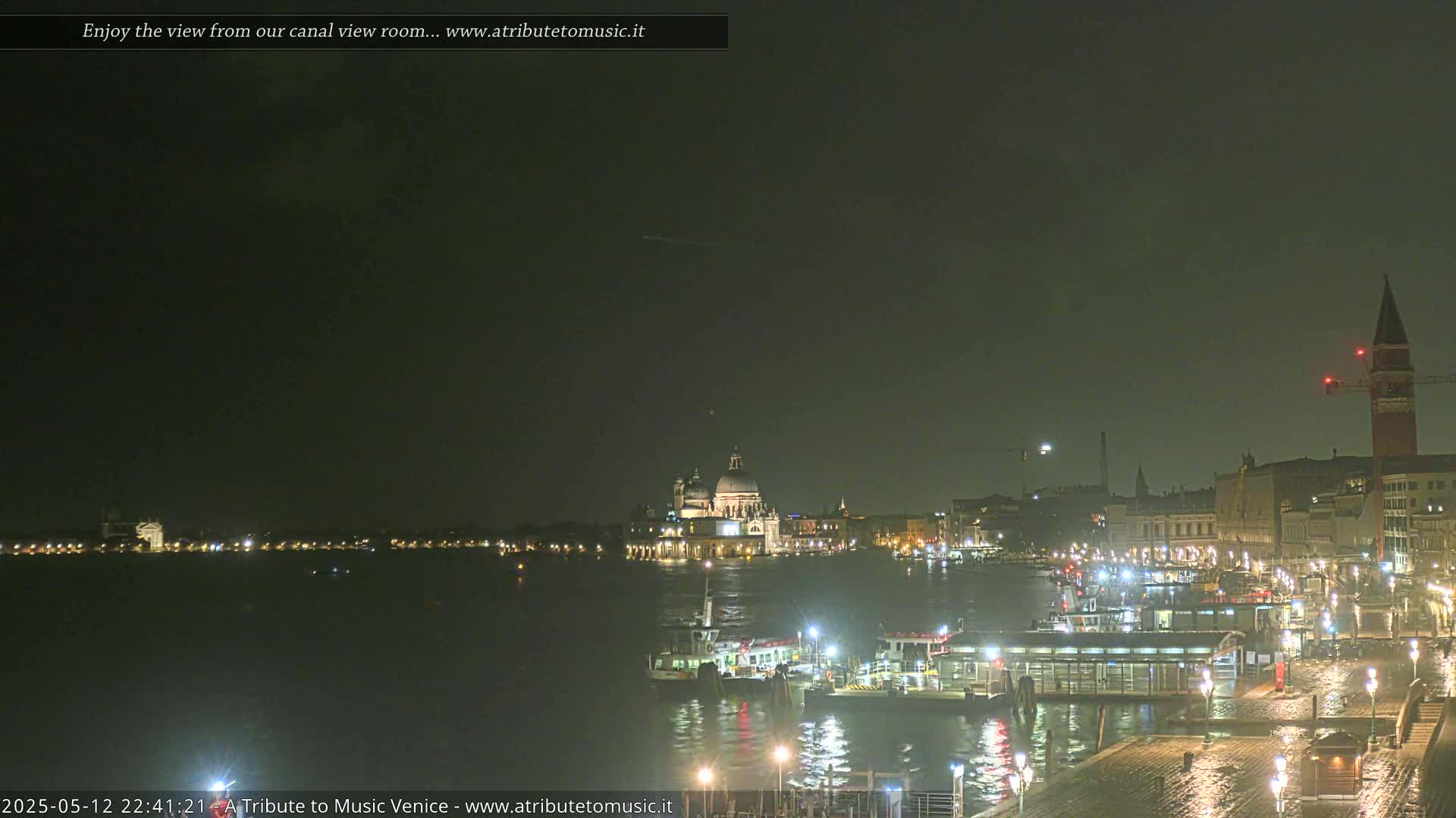 A nighttime view of a canal in Venice, showing boats moored at a dock with illuminated buildings and a church in the background, under a dark, possibly overcast sky.