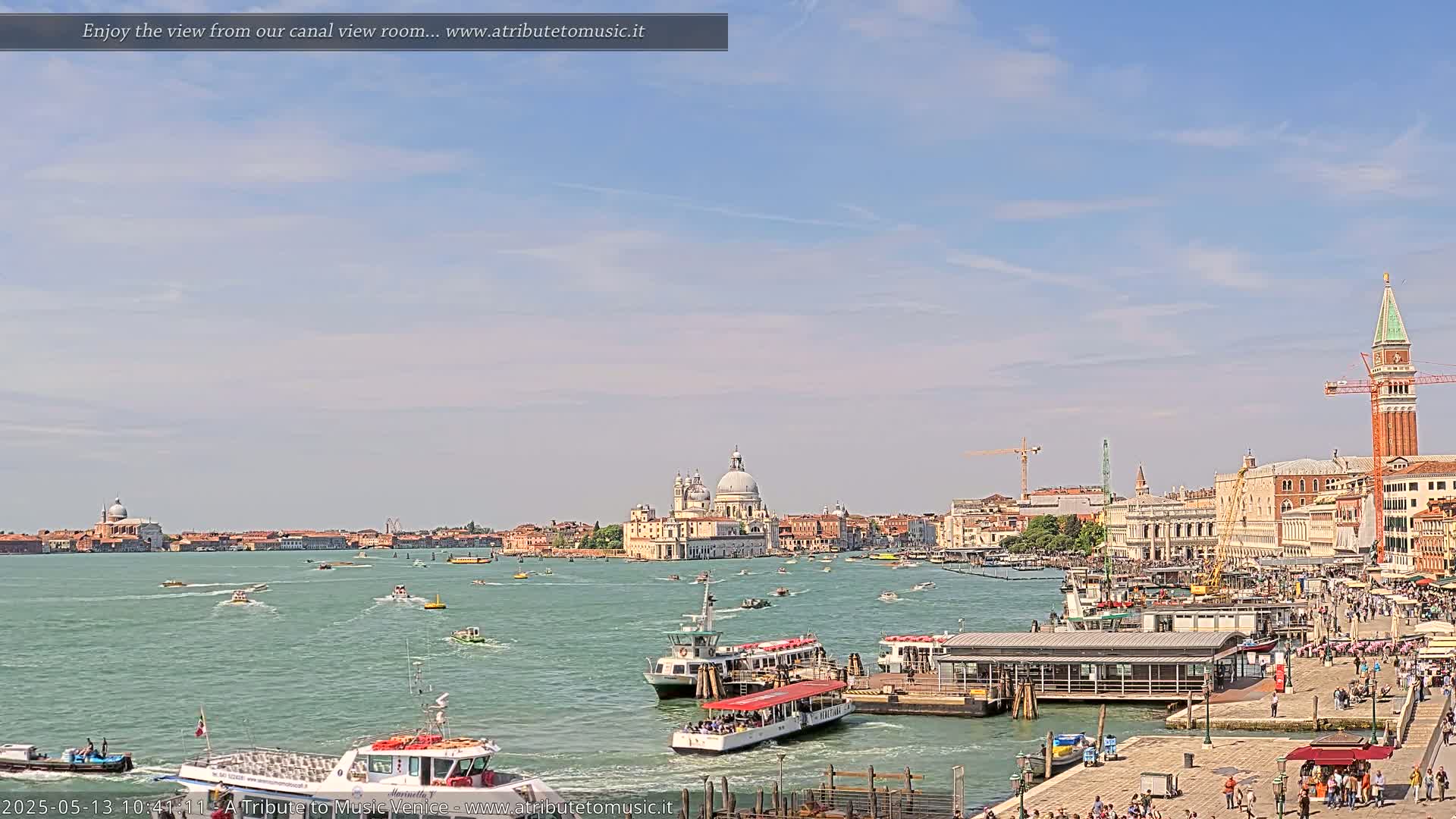 A sunny day in Venice shows a wide canal with numerous boats, a waterfront with buildings and people, and the Basilica di Santa Maria della Salute in the background.