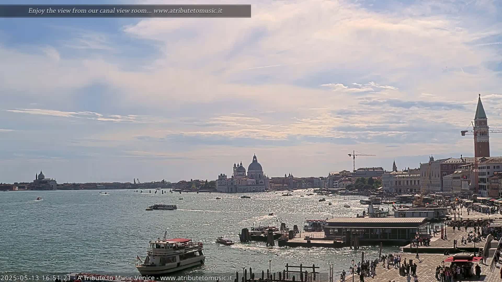 A sunny day in Venice shows a wide canal with numerous boats, a cityscape featuring a large domed church and a tall bell tower, and many people walking along a waterfront.
