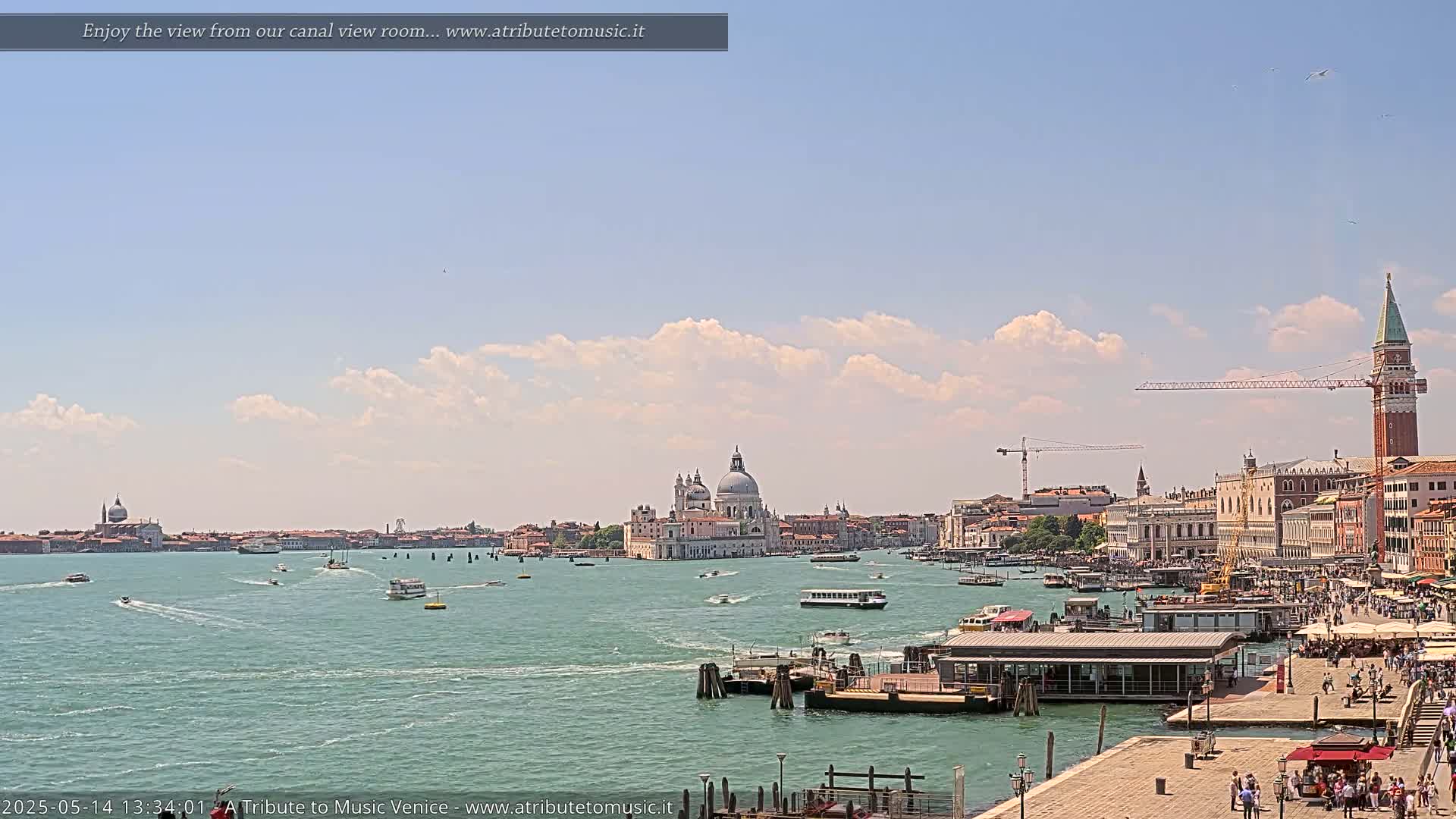 A sunny day in Venice shows a wide canal with boats, buildings, and a large church in the distance, under a partly cloudy sky.
