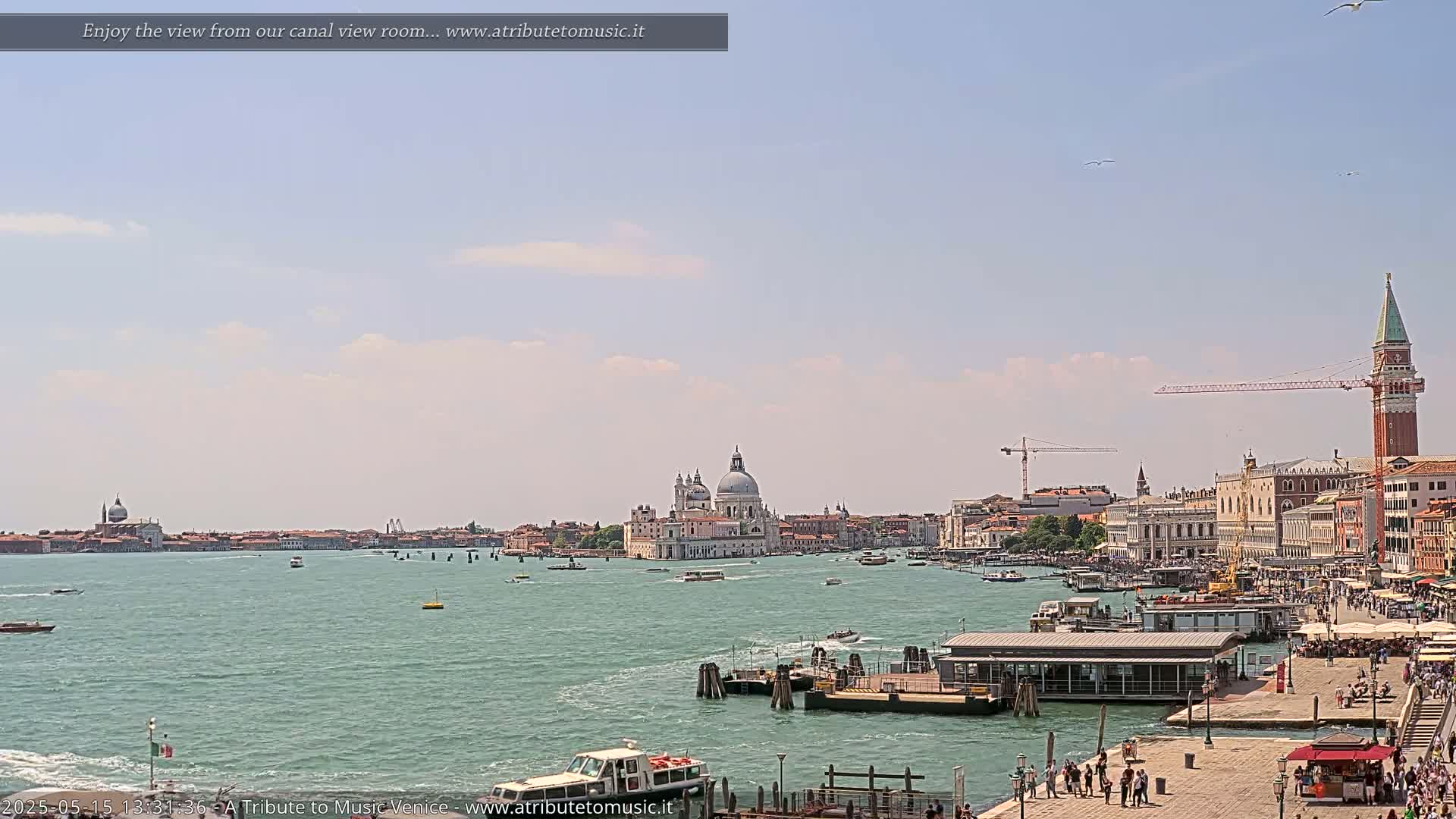 A wide shot of a Venetian waterway on a partly sunny day shows boats, buildings, and a bell tower along the shore.