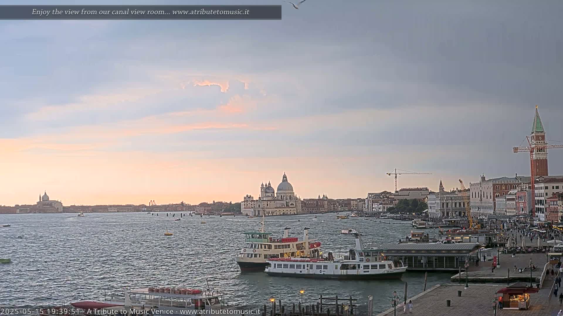 A tranquil Venetian canal scene at sunset shows several water taxis and boats near a waterfront, with the Basilica di Santa Maria della Salute and St. Mark's Square visible in the distance.