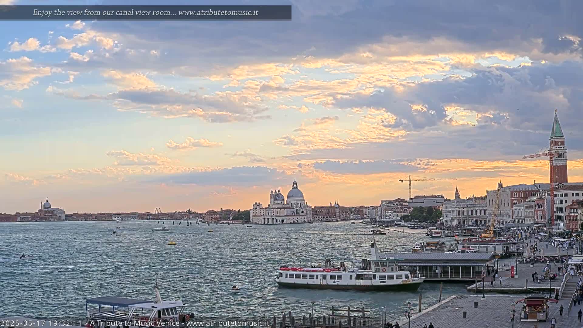A wide, calm canal in Venice, Italy, shows a cityscape at sunset under a partly cloudy sky.
