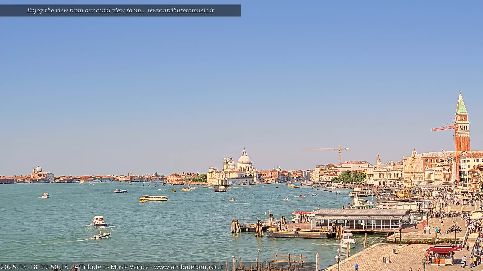 A sunny view of a Venetian canal, with boats and buildings, including a large church, lining the waterfront.