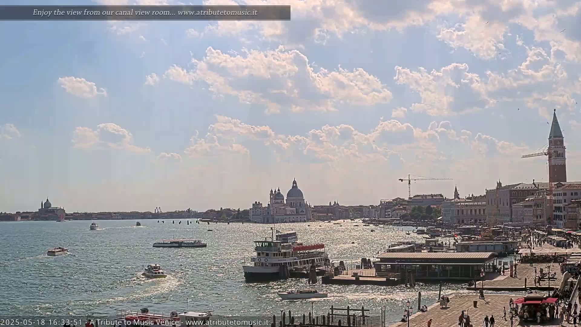 A sunny day in Venice shows a Venetian canal with boats, a cityscape including the Basilica di Santa Maria della Salute, and the St. Mark's Campanile in the distance under a partly cloudy sky.