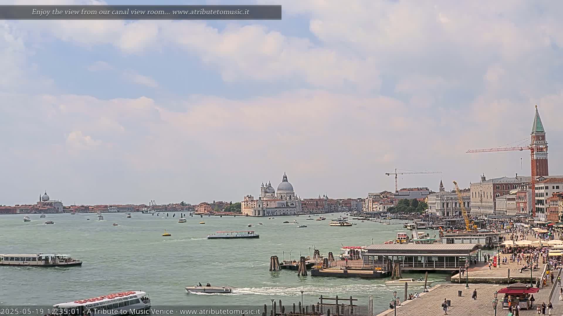 A partly cloudy sky overlooks a Venetian canal scene with numerous boats, buildings, and people gathered near a dock.