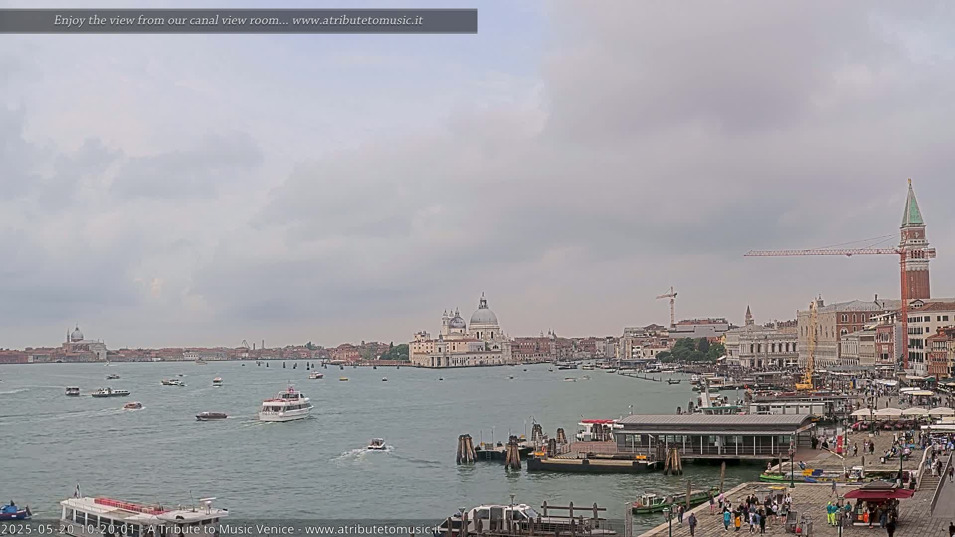 A wide, calm canal in Venice, under a cloudy sky, is filled with boats, and features a cityscape including a large domed building and a tall bell tower.