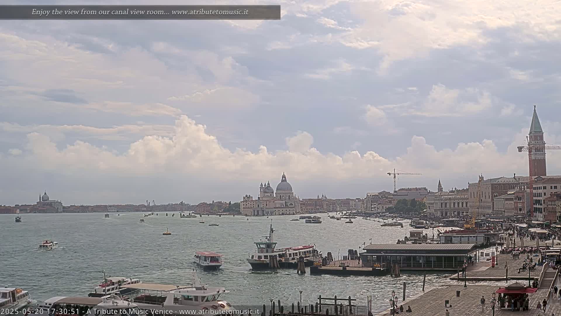 A wide shot of a Venetian canal shows several boats, a waterfront area with people, and buildings including a large church under a partly cloudy sky.