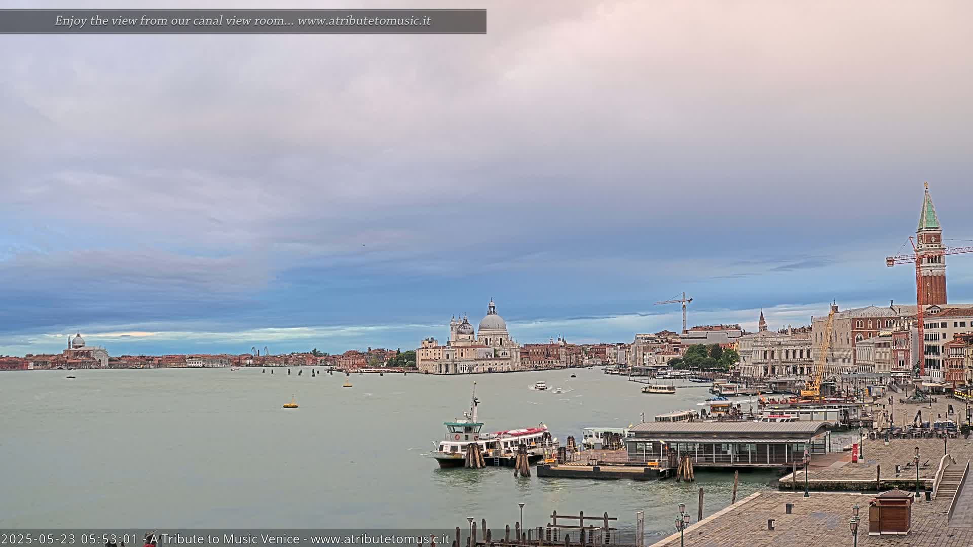 A wide view of a calm canal in Venice, under a mostly cloudy sky, shows a collection of buildings, boats, and a ferry terminal.