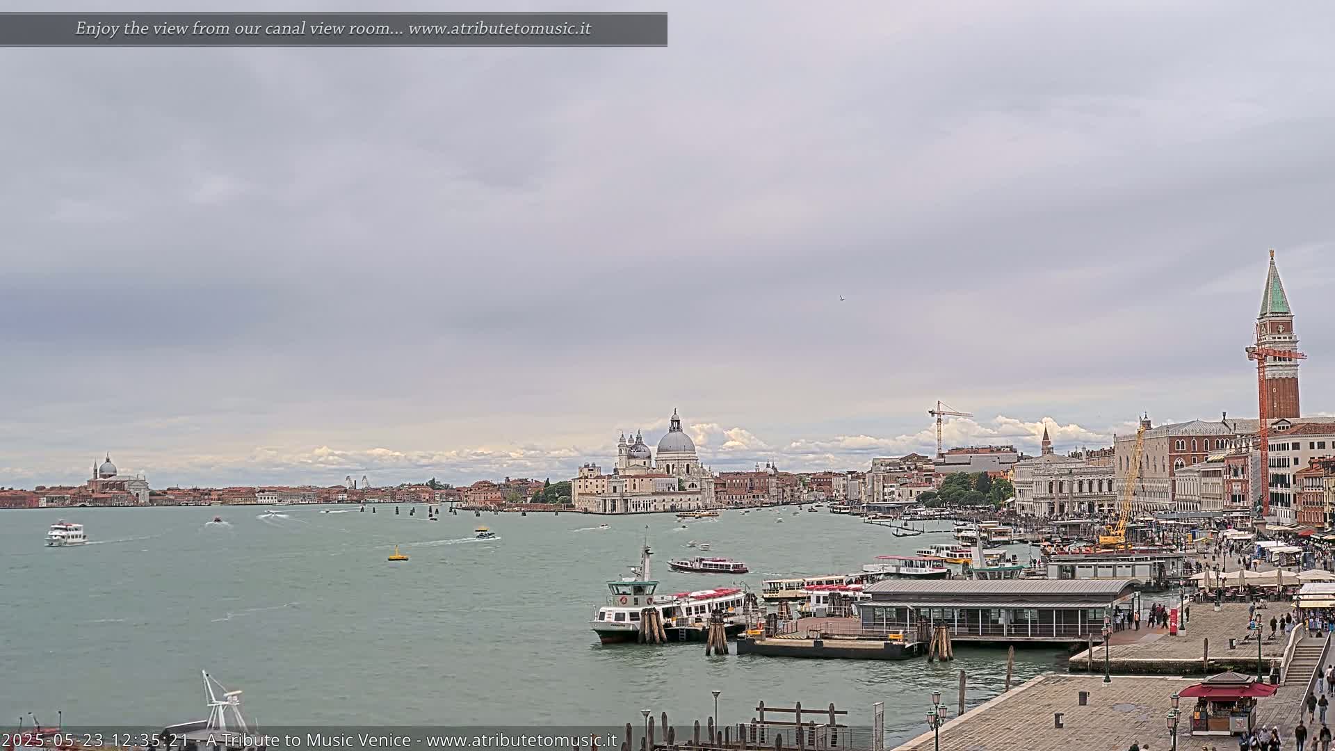 A wide shot of a Venetian canal on an overcast day shows several boats near a dock, with the Basilica di Santa Maria della Salute and other Venetian buildings in the background.