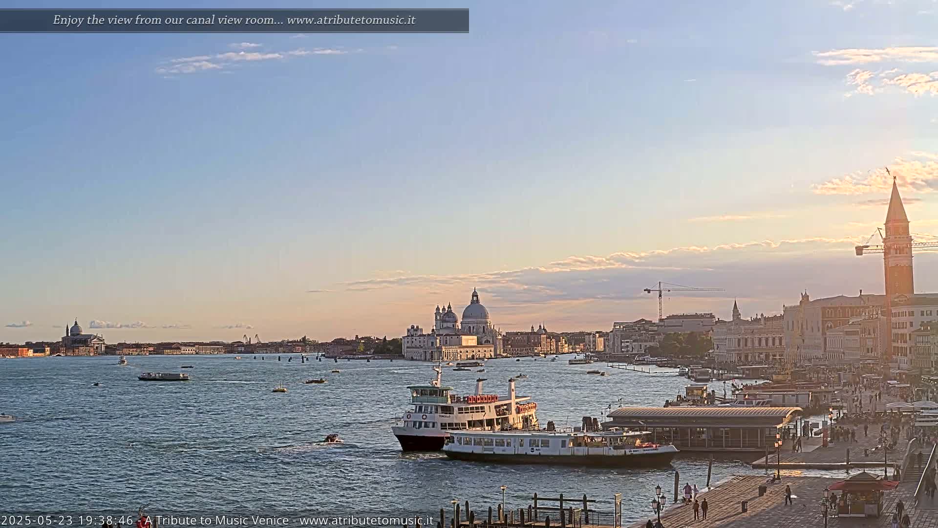 A wide canal view of Venice at sunset, showing a ferry docked near a waterfront, with boats on the water and a cityscape including the Basilica di Santa Maria della Salute in the background under a mostly clear sky.