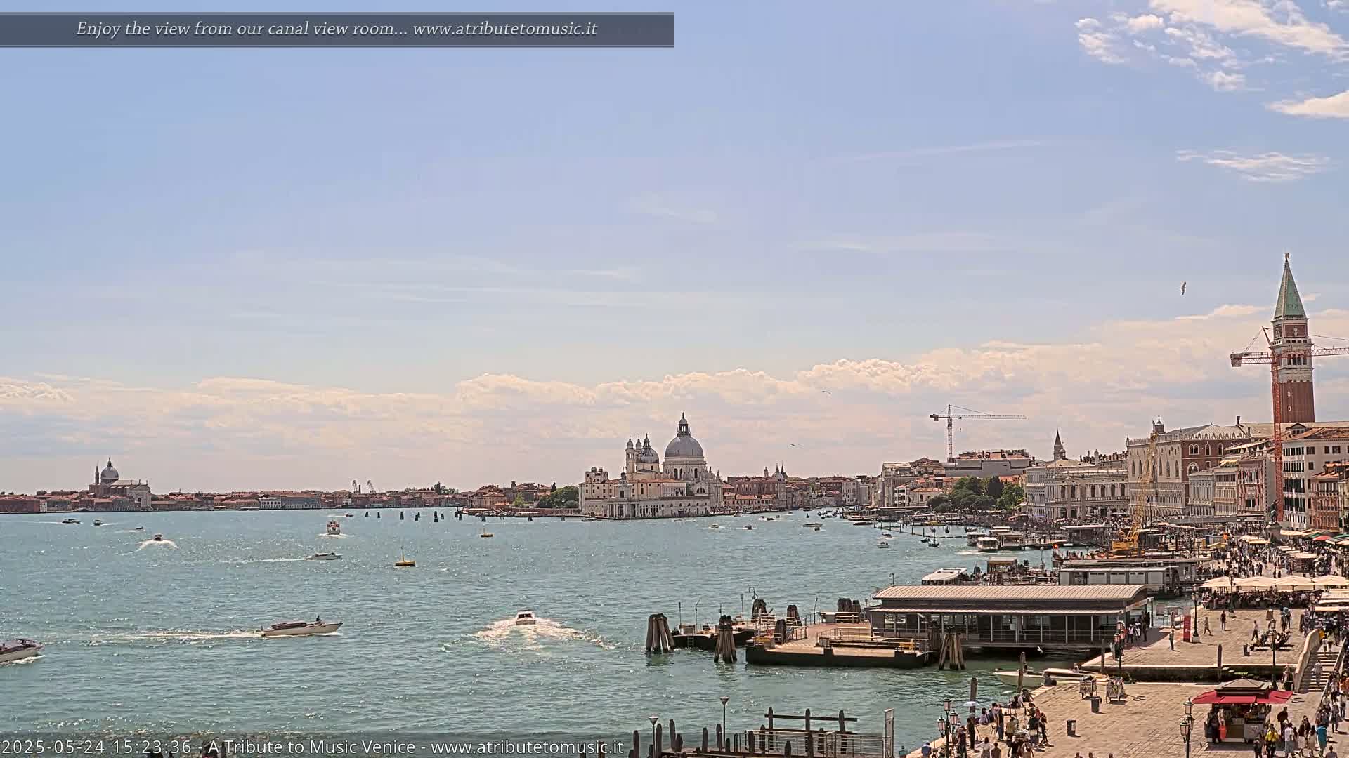 A wide shot of a Venetian canal shows boats on the water, buildings and a church along the far shore, and a paved area with people walking in the foreground under a partly cloudy sky.