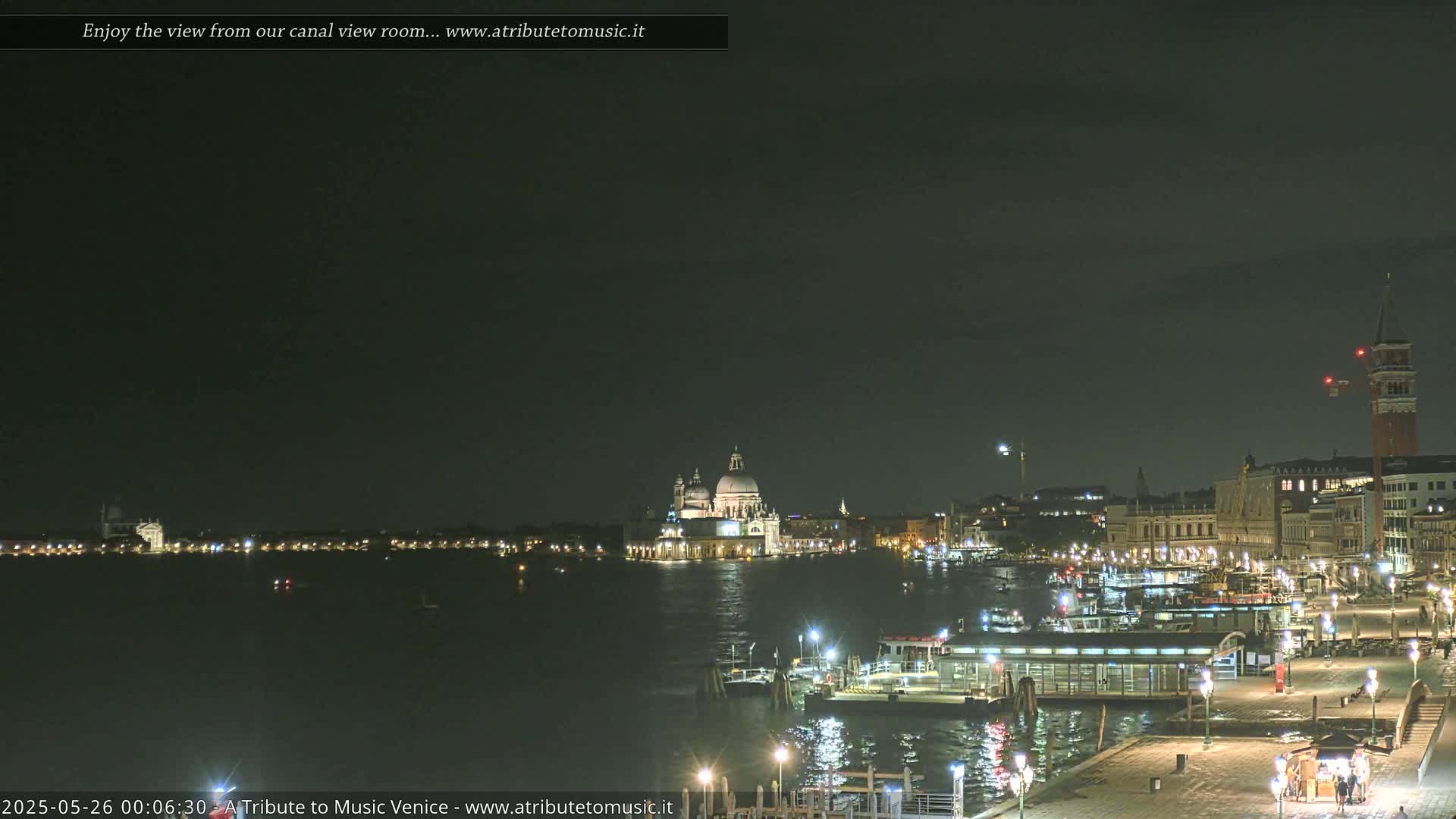 A nighttime view across a calm canal shows illuminated buildings and boats in Venice under a dark sky.
