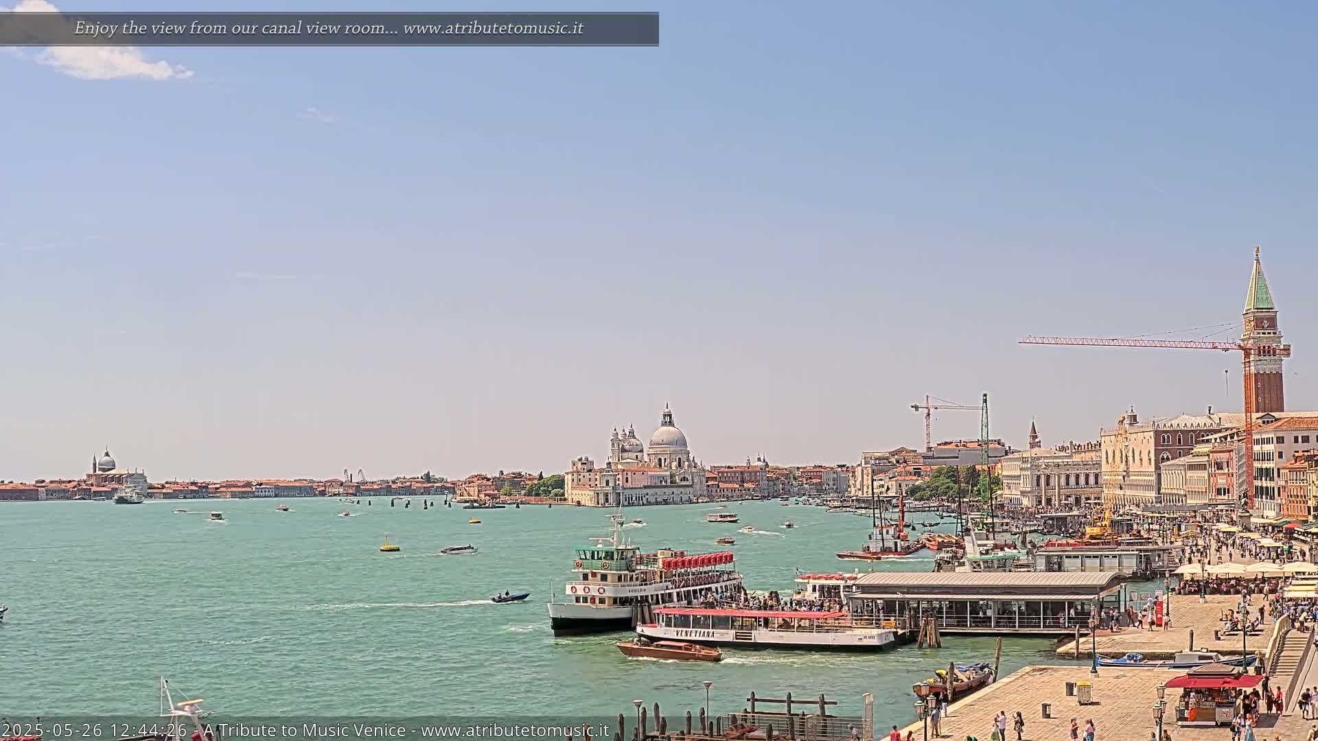 A sunny day in Venice shows a wide canal with several boats, including passenger ferries, near a waterfront bustling with people and buildings, including a large church with a dome.