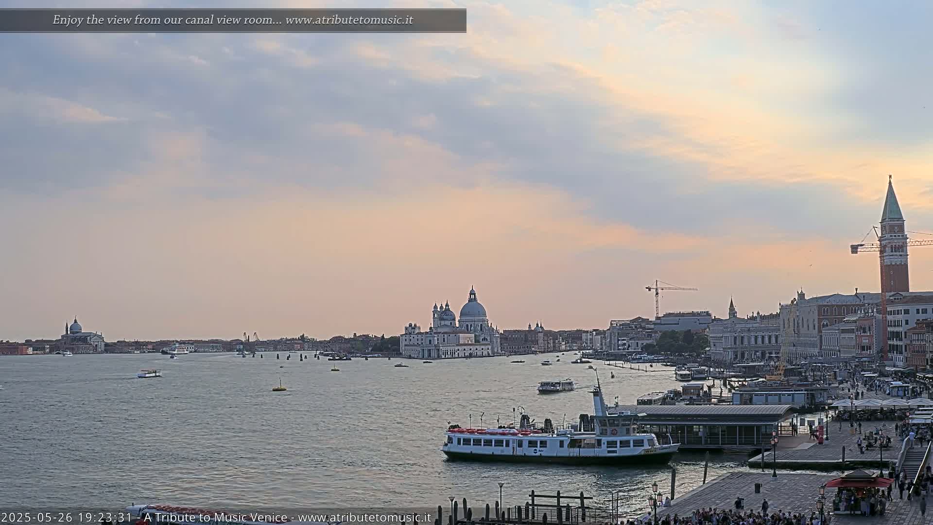 A wide canal scene at sunset shows a Venetian cityscape with boats on the water, a large church in the distance, and people gathered along the waterfront.