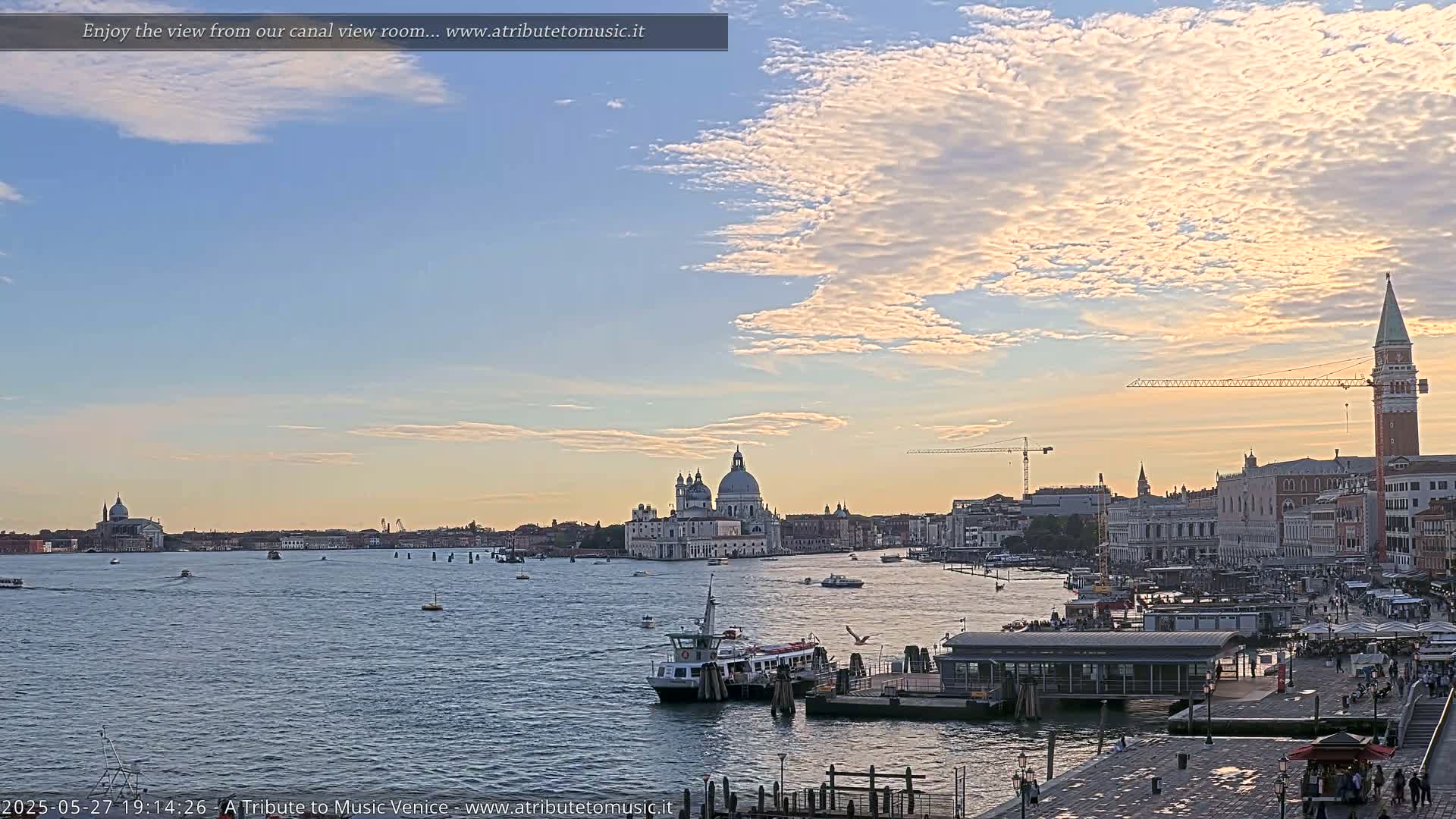 A wide canal view of Venice at sunset, under a partly cloudy sky, shows a ferry boat and numerous buildings along the waterfront, including a large domed church.