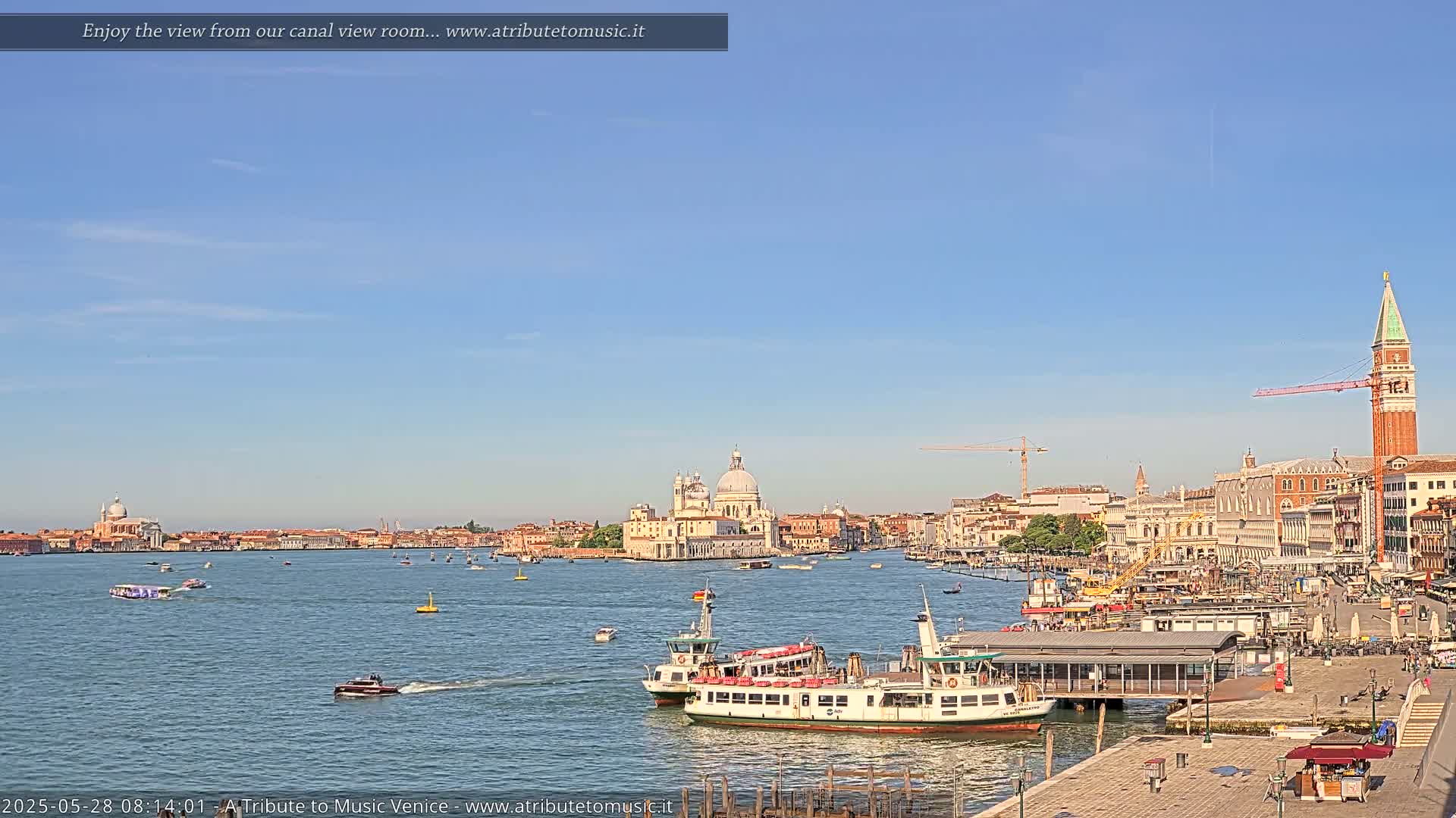 A clear blue sky overlooks a Venetian waterway with several boats, including waterbuses, and a cityscape featuring a prominent church and the St. Mark's Campanile.