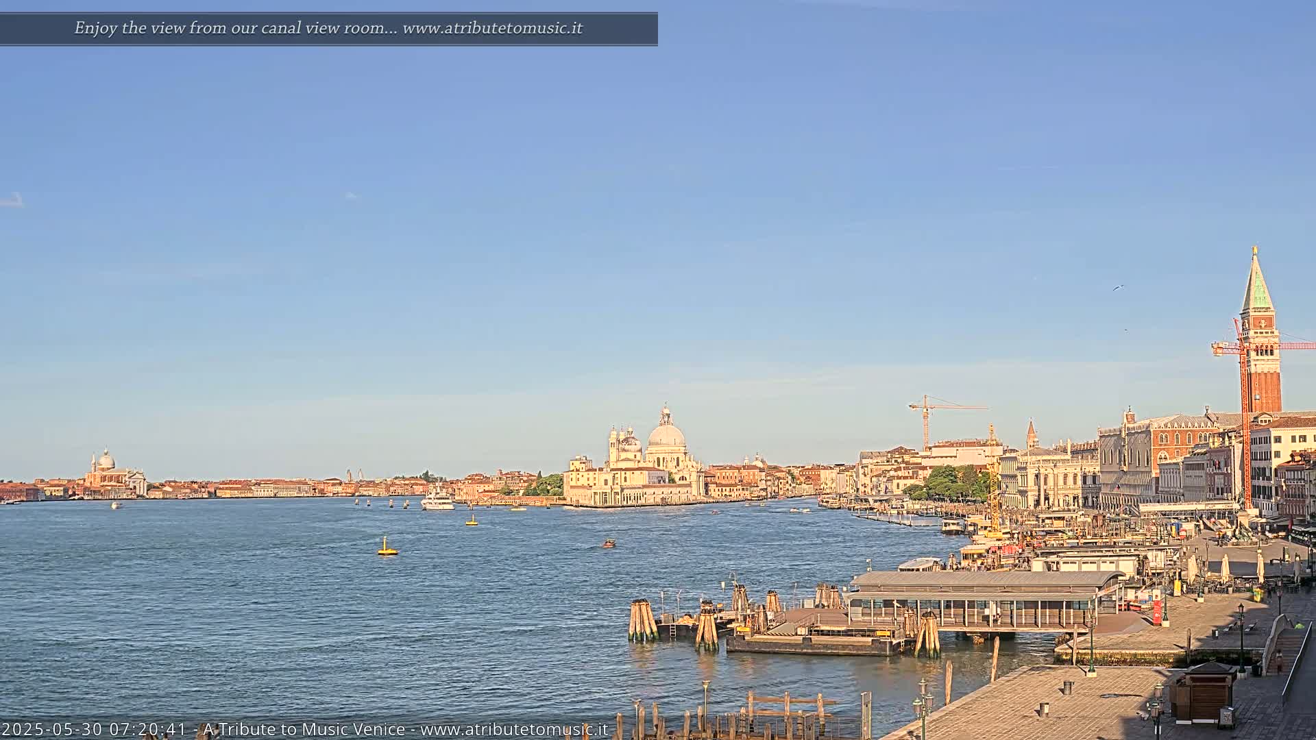 A wide shot of a calm Venetian canal on a clear, sunny day, shows a cityscape with several buildings and a church in the distance, along with a water taxi station in the foreground.