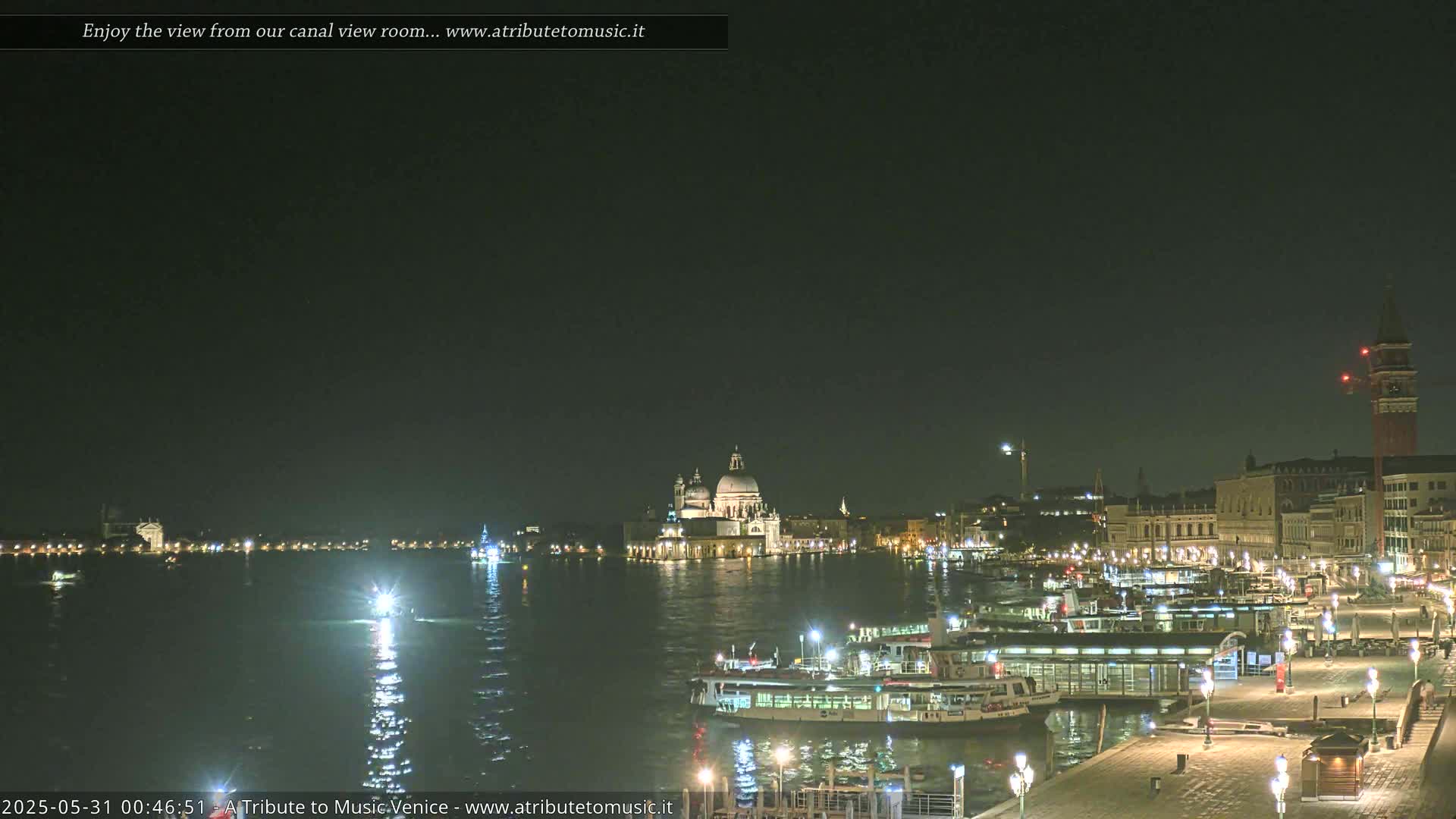 A nighttime view of a canal in Venice, showing illuminated buildings, boats, and the Basilica di Santa Maria della Salute.