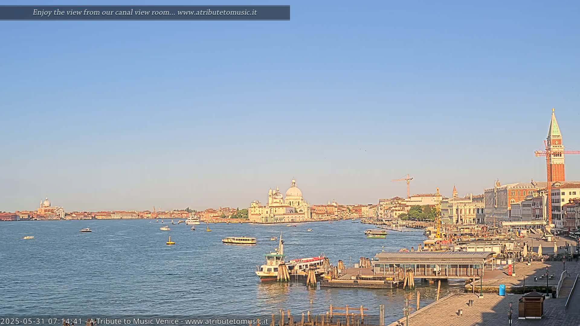 A calm canal scene in Venice shows a clear blue sky, several boats on the water, and the city's buildings, including a large church and the St. Mark's Campanile, along the shore.