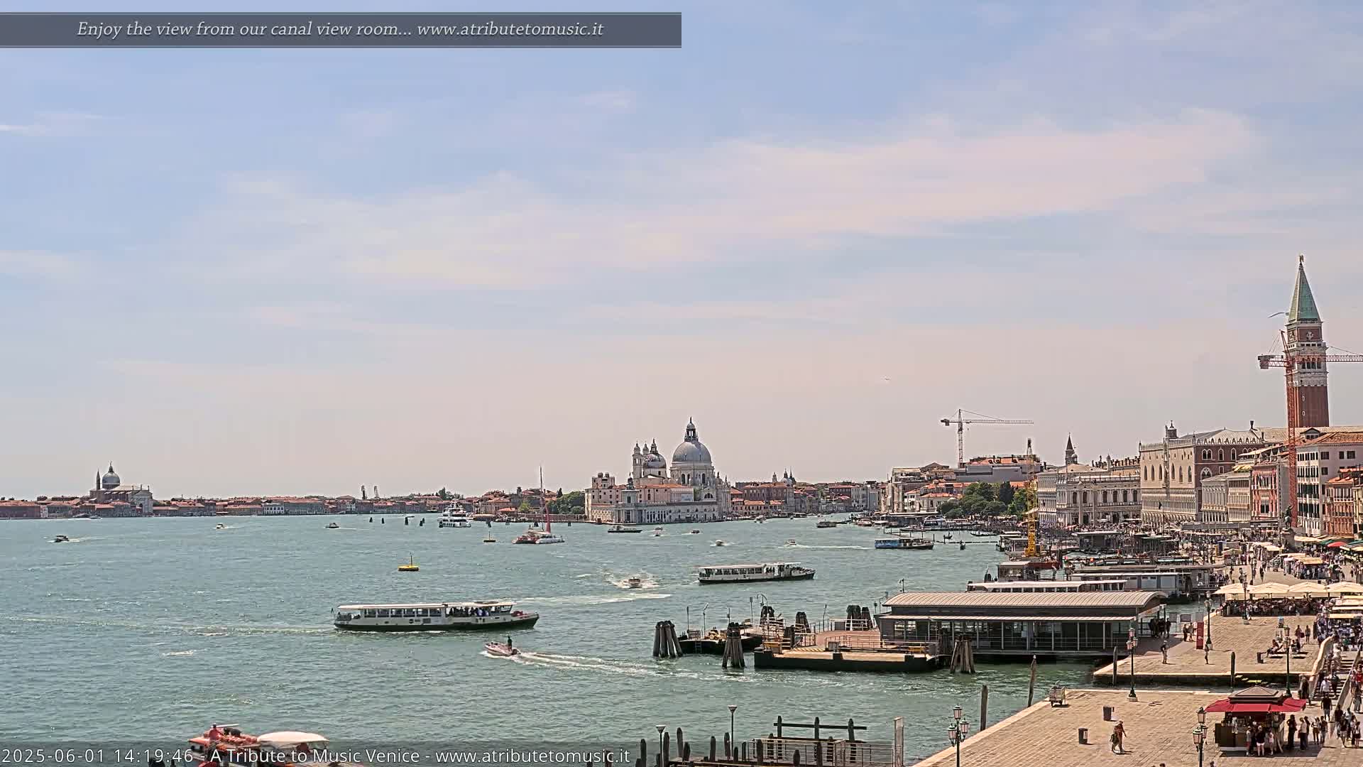 A sunny day in Venice shows a wide canal with numerous boats, a cityscape including the Basilica di Santa Maria della Salute, and a waterfront area with people.