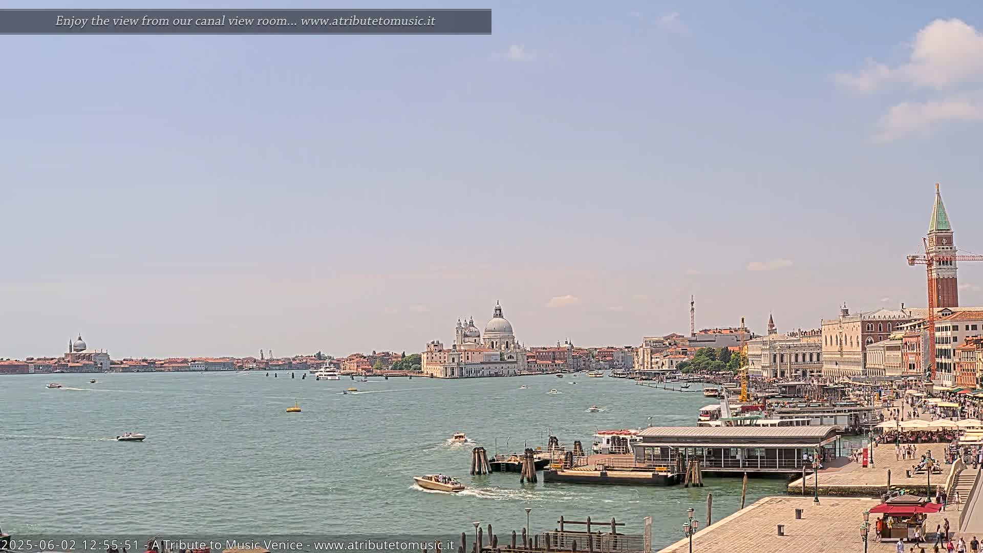A sunny day in Venice shows a wide canal with boats, a dock, and a cityscape including the Basilica di Santa Maria della Salute.