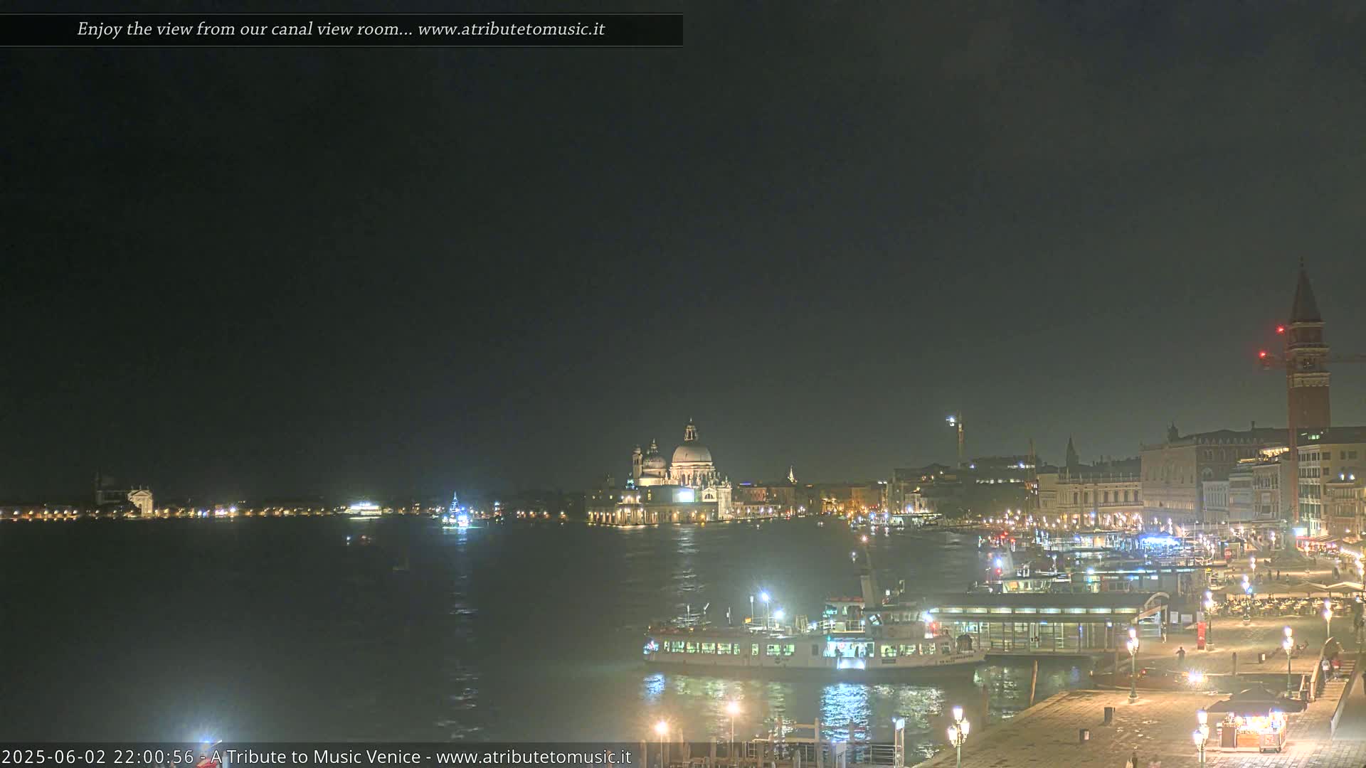 A nighttime view across a calm canal shows illuminated buildings and boats in Venice, under a dark sky.