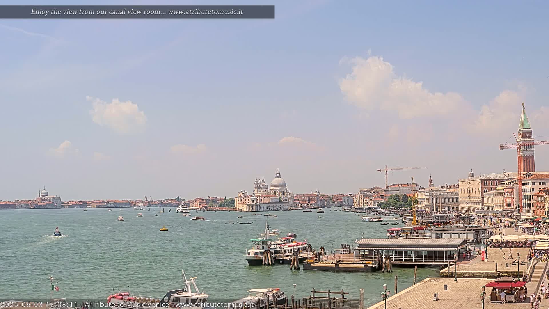 A sunny day in Venice shows a wide canal with numerous boats, buildings along the shore including a large domed church, and a tall bell tower in the distance.