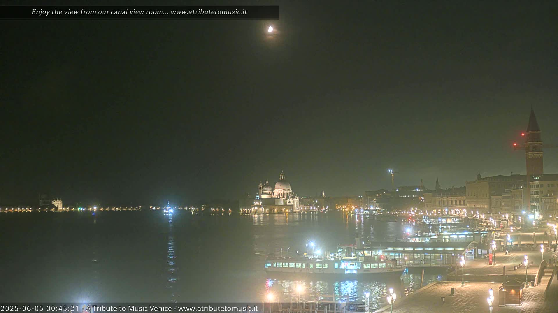 A nighttime view of a city waterfront with boats, buildings, and a large domed structure under a dimly lit sky, showing a partially visible moon.