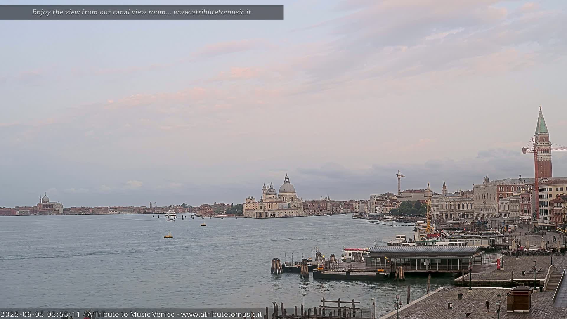 A wide shot shows a calm canal in Venice, under an overcast sky, with boats and buildings, including a large church, visible in the distance.