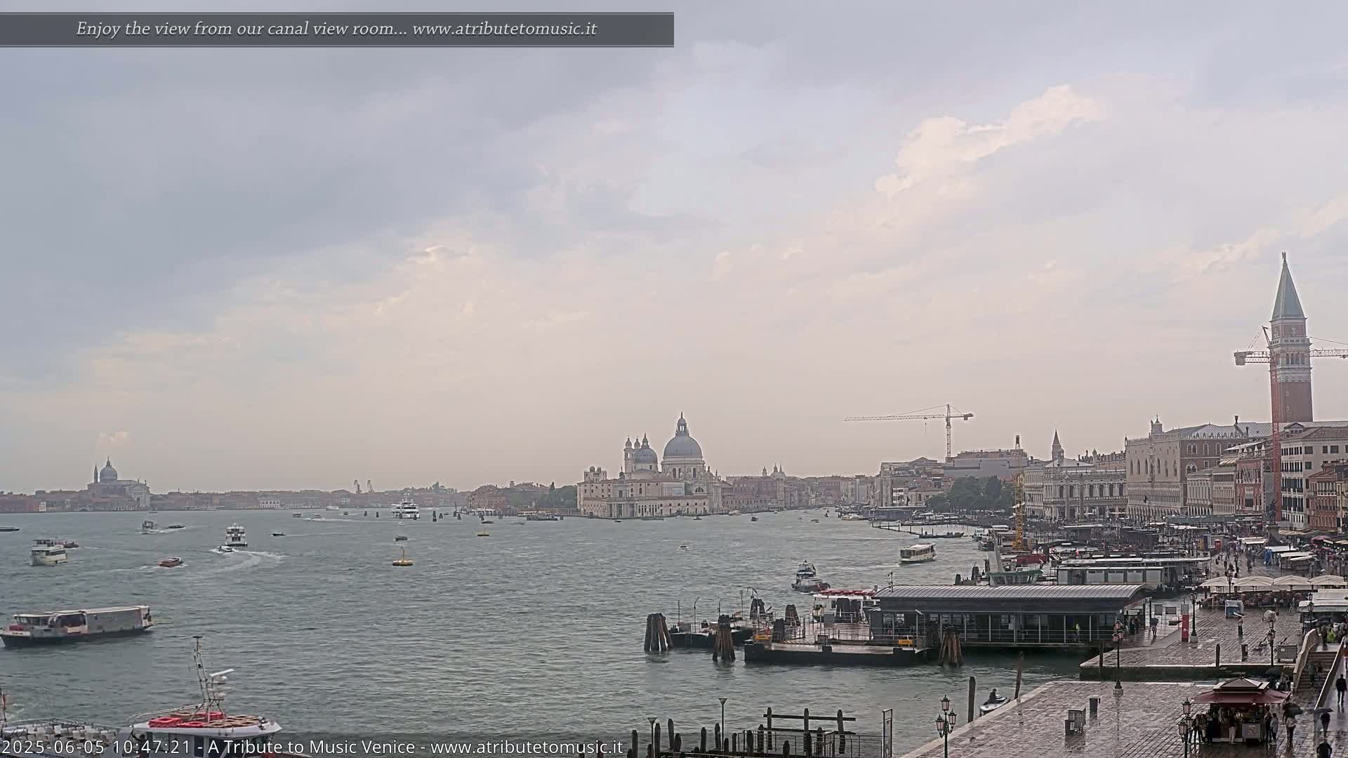 A wide shot of a waterway in Venice, Italy, on an overcast day, shows numerous boats, buildings, and a large church across the water, with some people visible near a dock in the foreground.