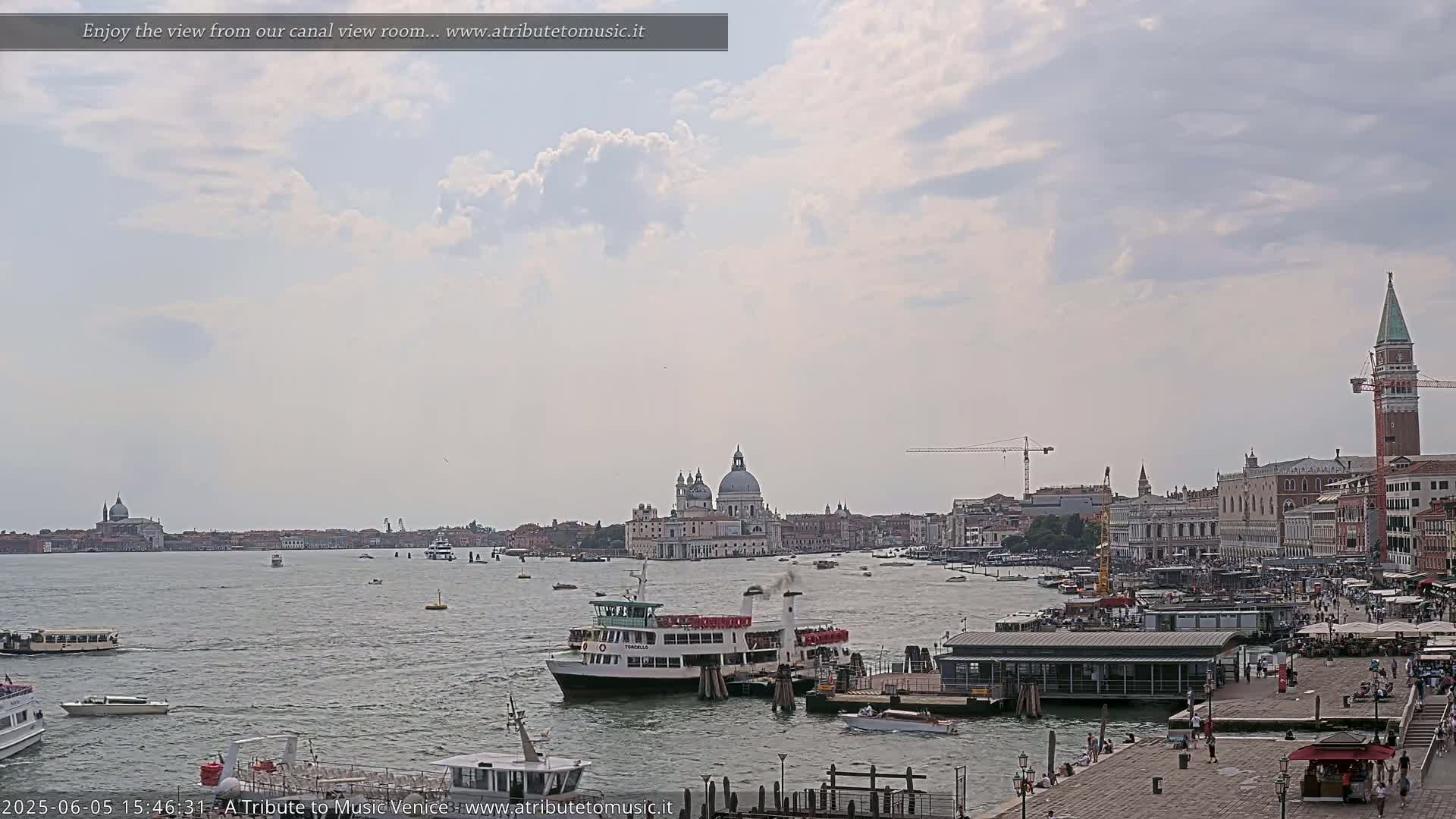 A wide shot of a Venetian canal on a partly cloudy day shows several boats, a ferry, and the city skyline including a large church and a tall tower.