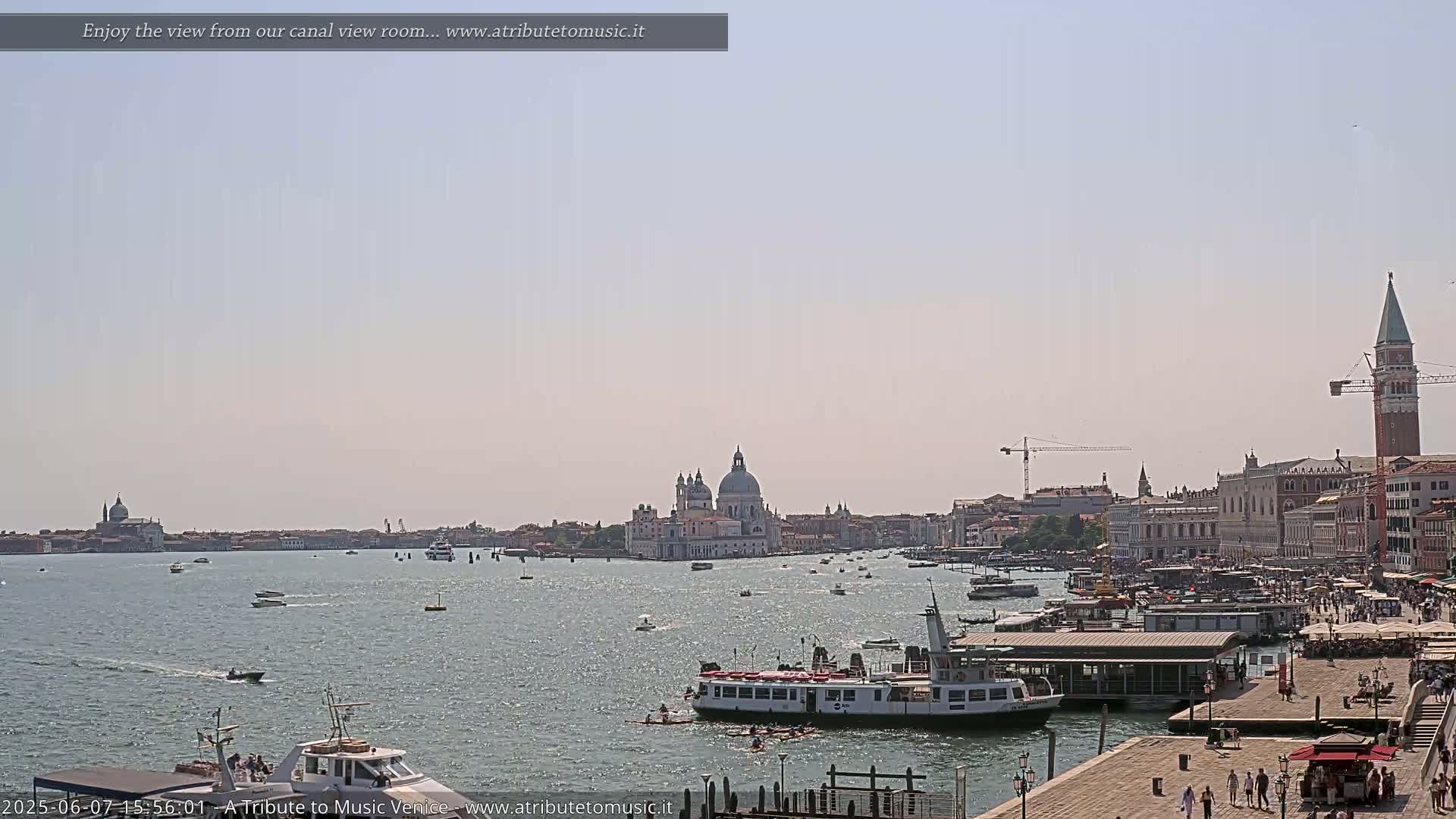 A wide shot of a Venetian canal on a sunny day shows numerous boats, a ferry, and a cityscape including a large domed church and a bell tower.