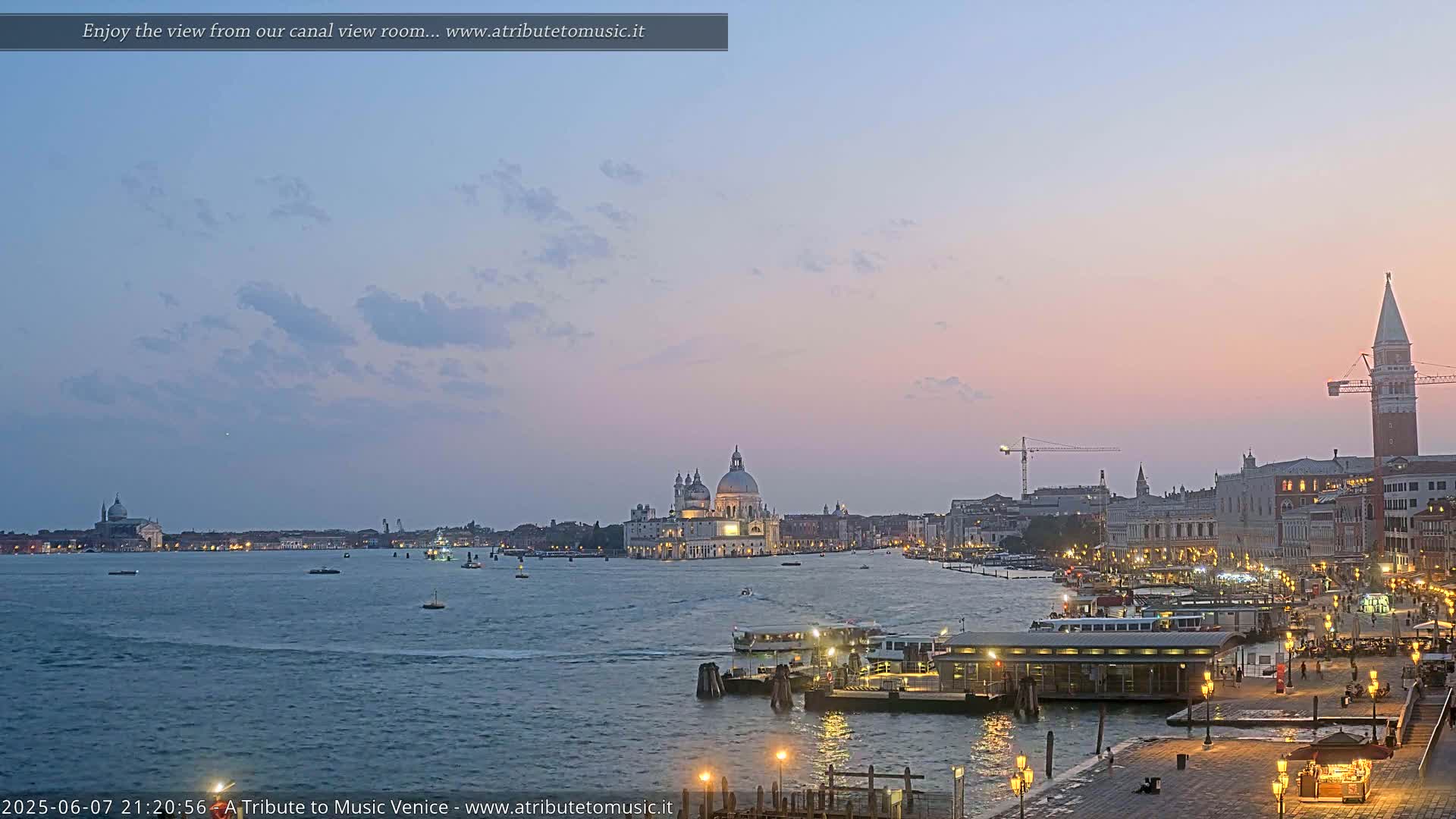 A twilight view across a calm canal shows the illuminated Venetian skyline, featuring a large domed church and a tall bell tower.