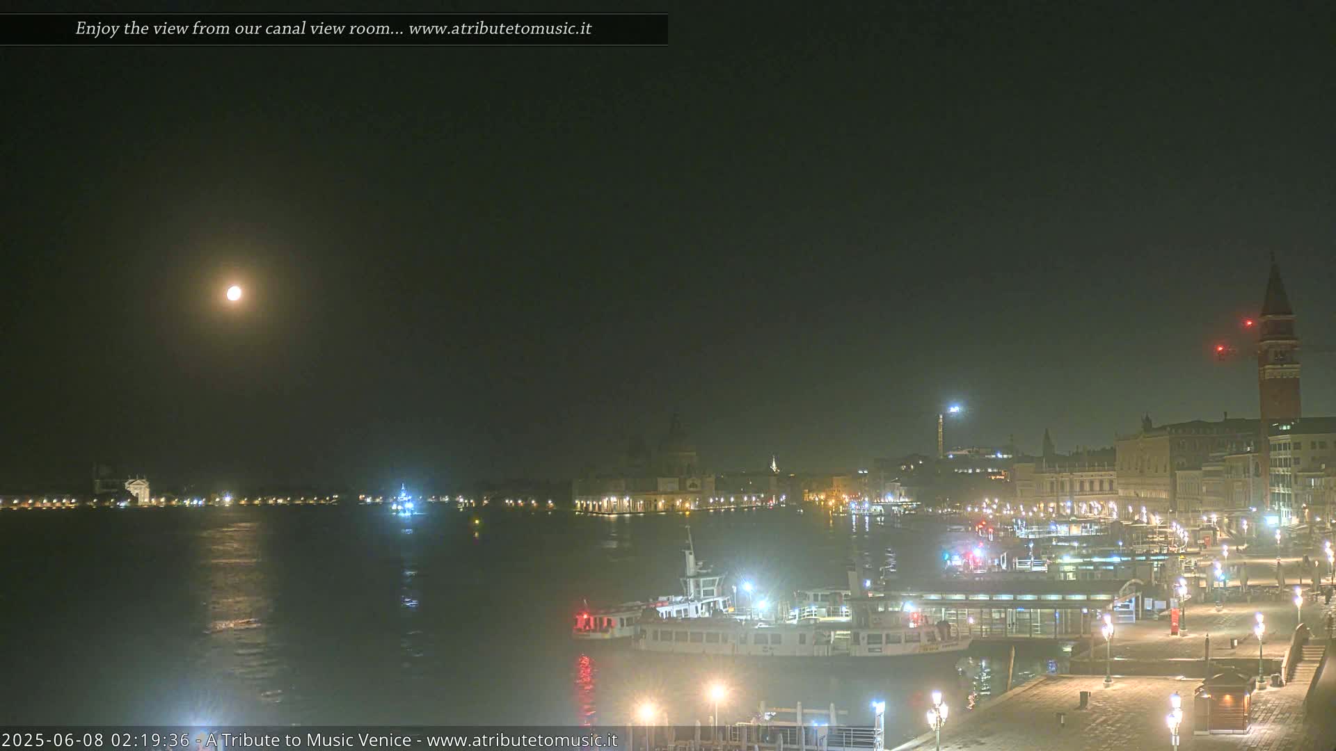 A nighttime view of a calm canal, with boats moored along a waterfront lit by streetlights, under a full moon in a hazy sky.