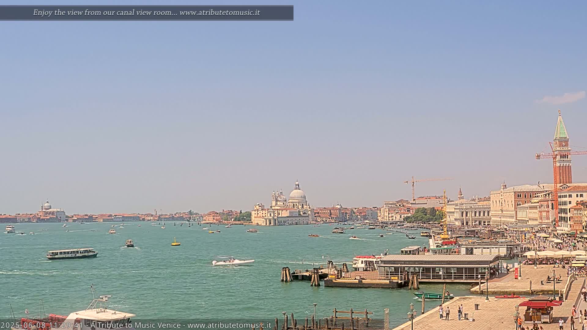 A sunny Venetian canal scene shows numerous boats and buildings, including a large basilica and a tall bell tower, under a clear blue sky.