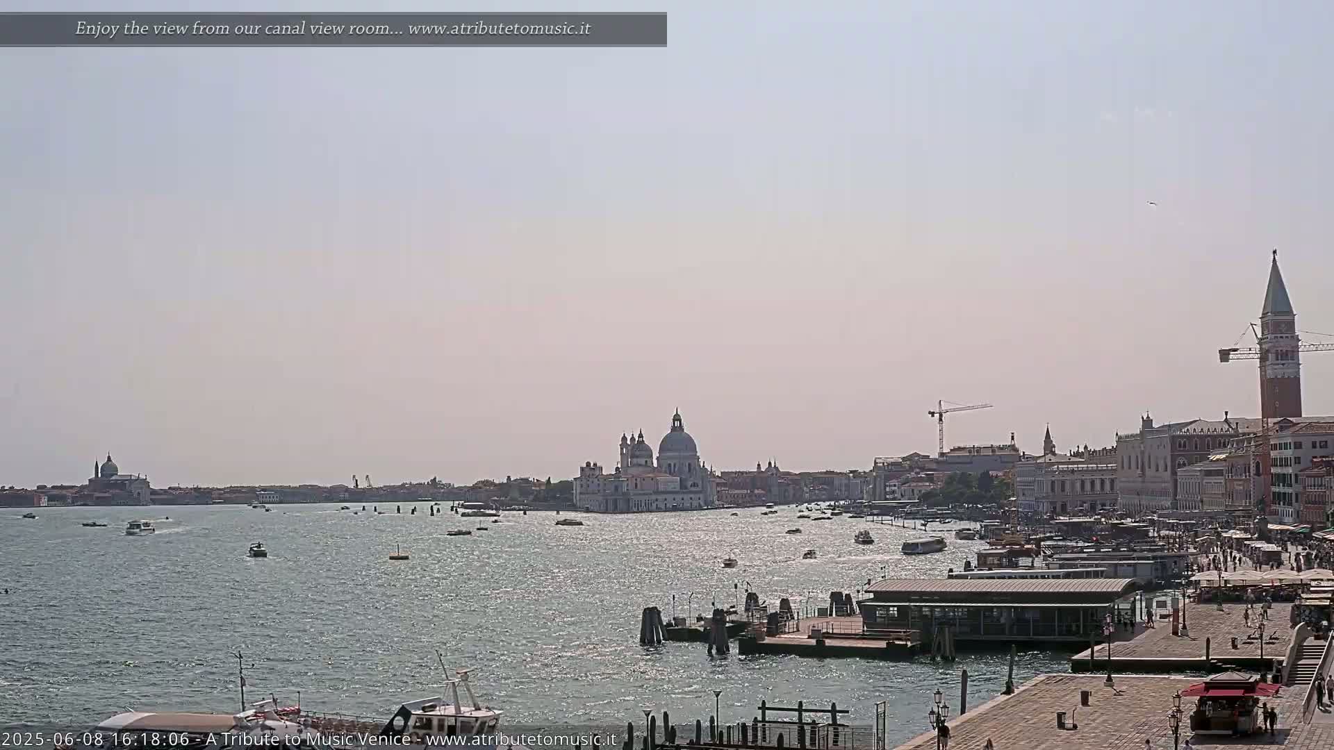 A sunny day in Venice shows a canal with numerous boats, a waterfront with buildings and people, and the Basilica di Santa Maria della Salute in the distance.