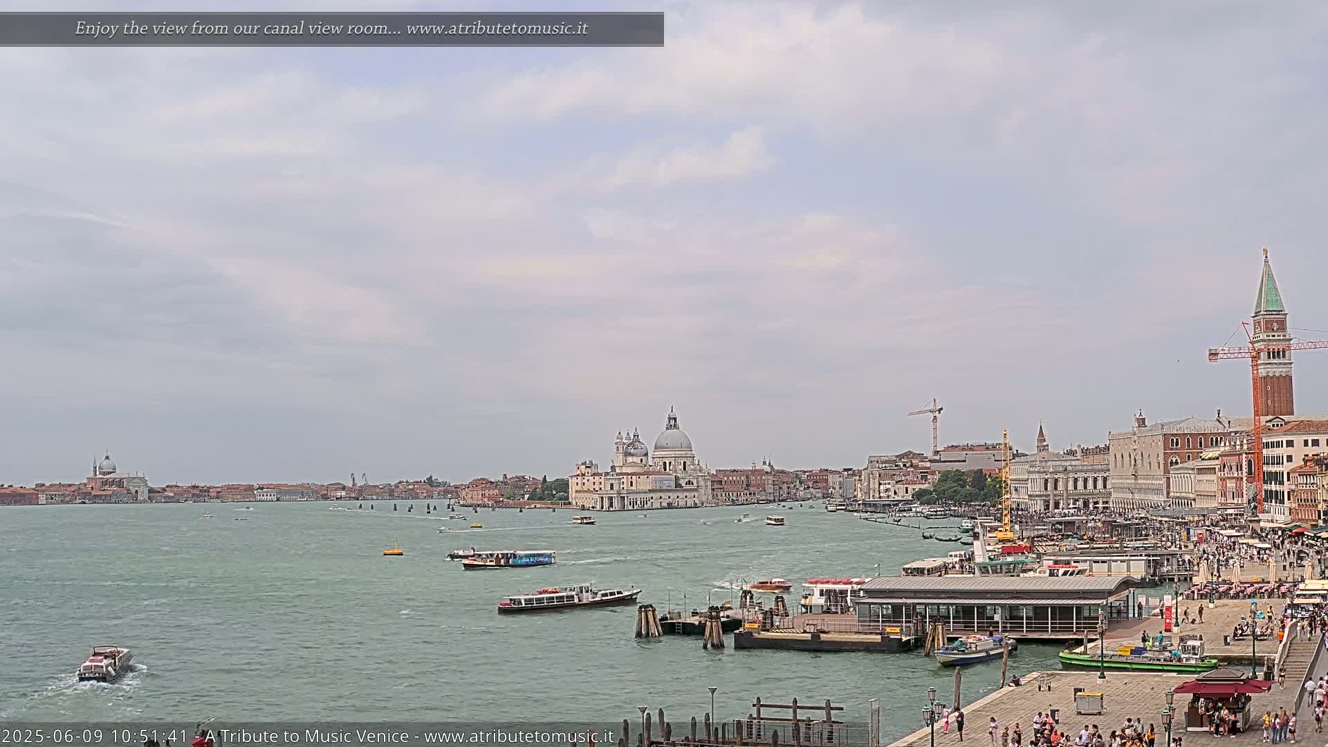 A wide, calm canal in Venice, Italy, is dotted with boats under a lightly cloudy sky, with several large buildings and a tall tower visible on the far shore.