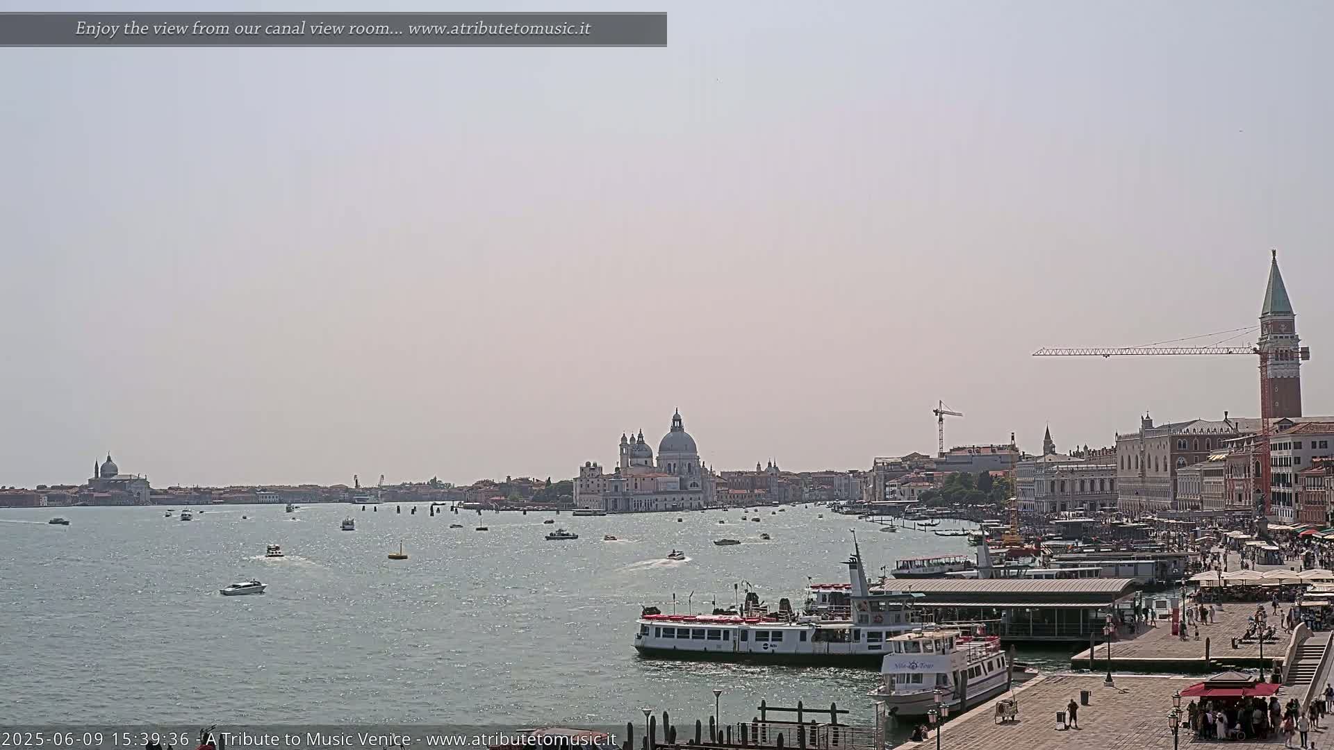 A hazy, sunny day reveals a Venetian canal scene with numerous boats, a waterfront cityscape, and the iconic Basilica di Santa Maria della Salute in the distance.