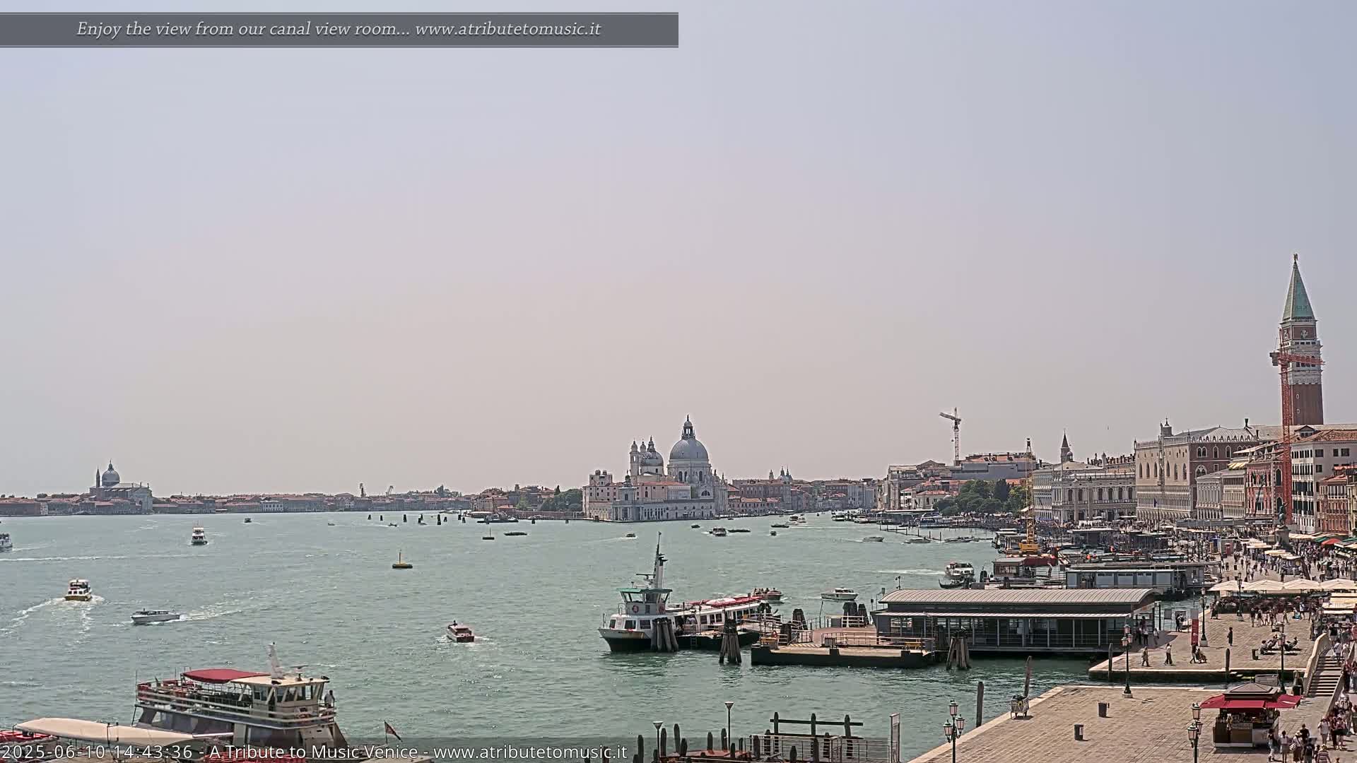 A sunny day in Venice shows a wide canal with numerous boats and buildings lining the waterway, including a large domed church in the distance.
