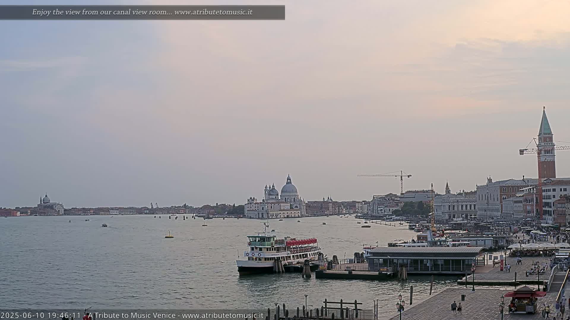 A ferry is docked near a waterfront building in Venice, Italy, under a pale, overcast sky, with the Basilica di Santa Maria della Salute visible in the distance.