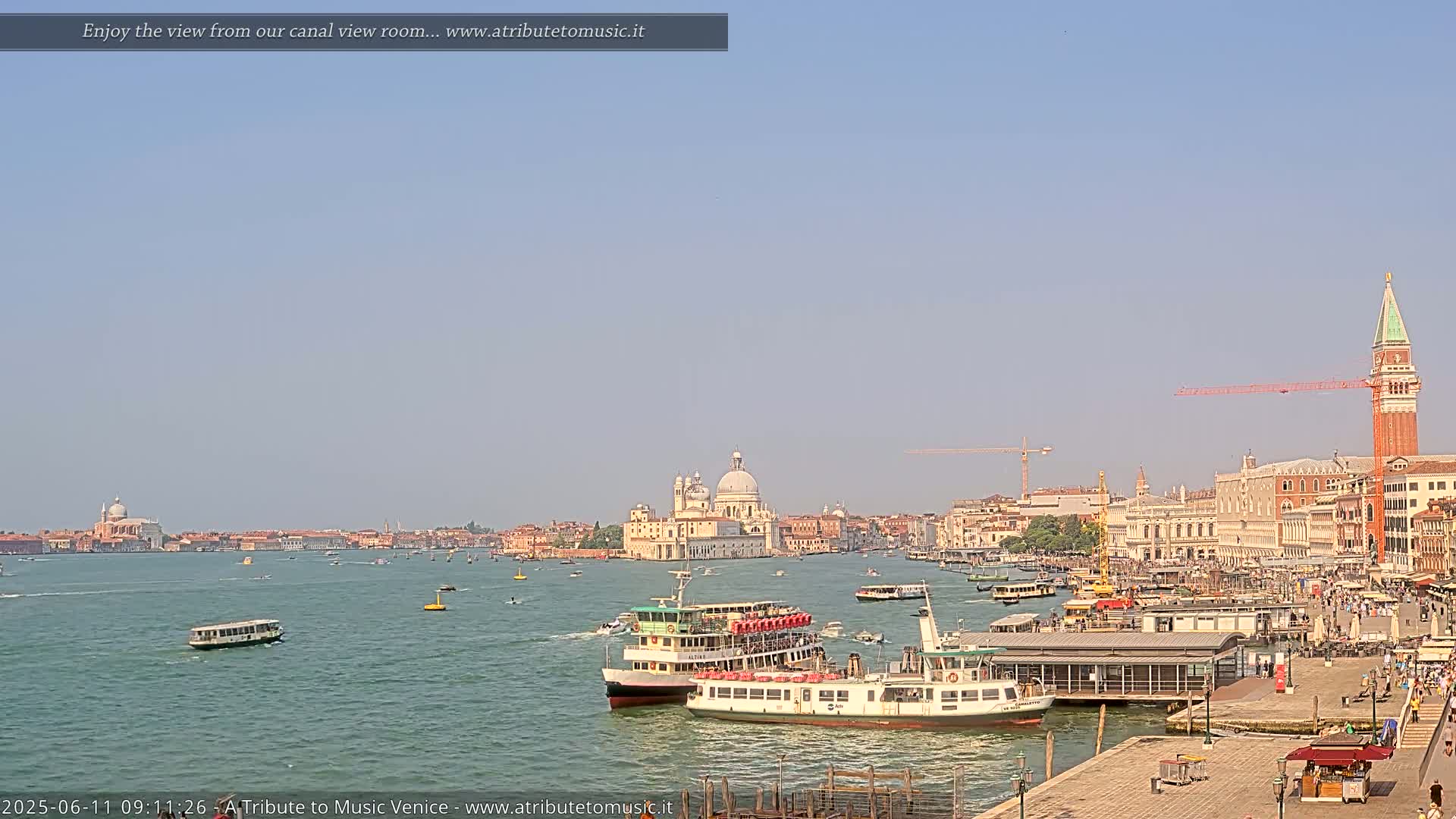 A wide shot of a Venetian canal on a clear, sunny day shows several boats and the city skyline in the distance, including a large white church.