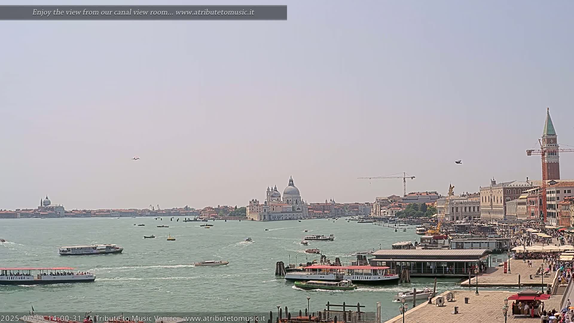 A sunny view of a Venetian canal shows numerous boats, buildings including a large domed church, and a tall bell tower, with two small aircraft visible in the sky.