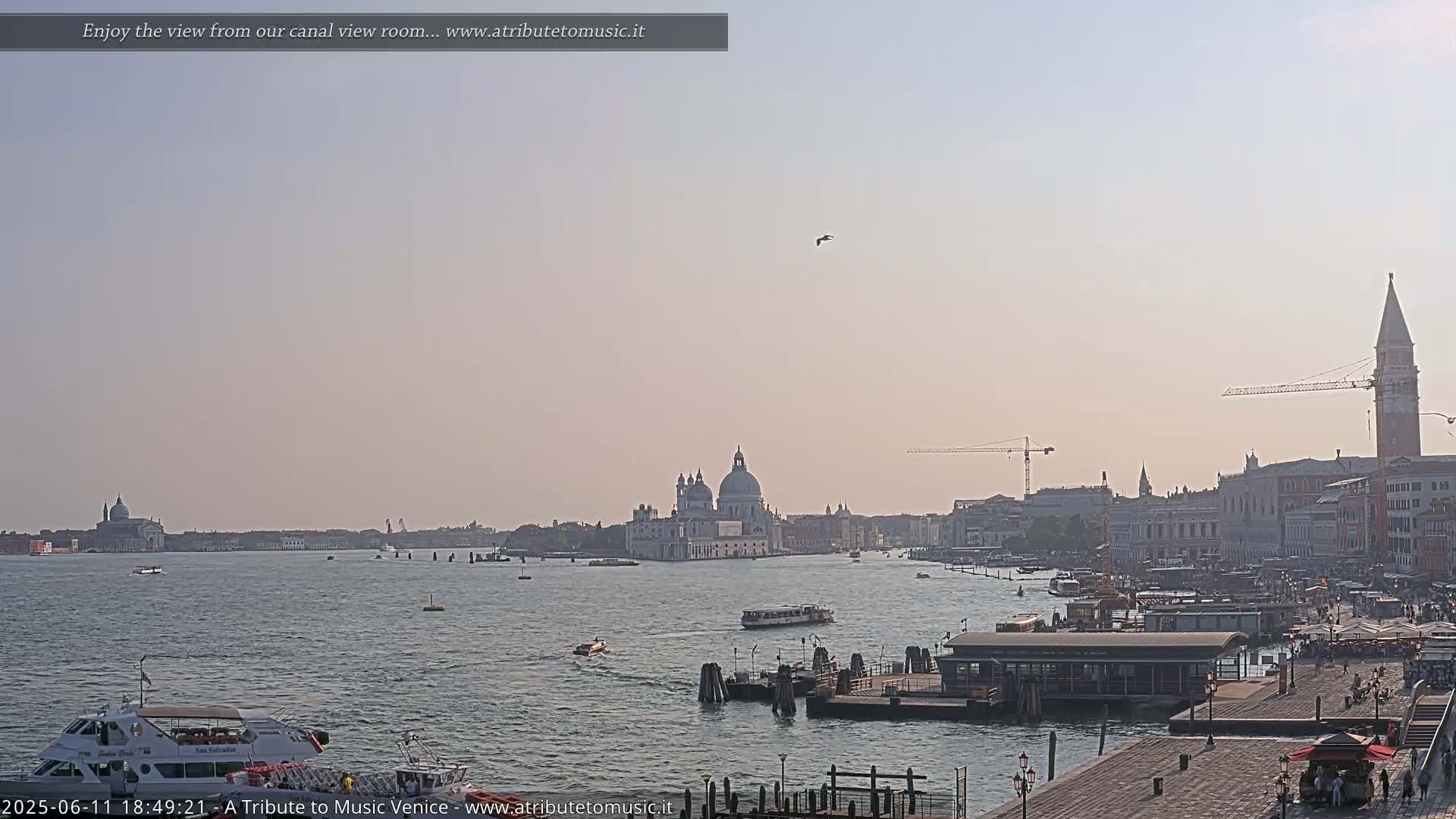 A hazy, sunny view of a Venetian canal shows boats, buildings, and a large church in the distance.
